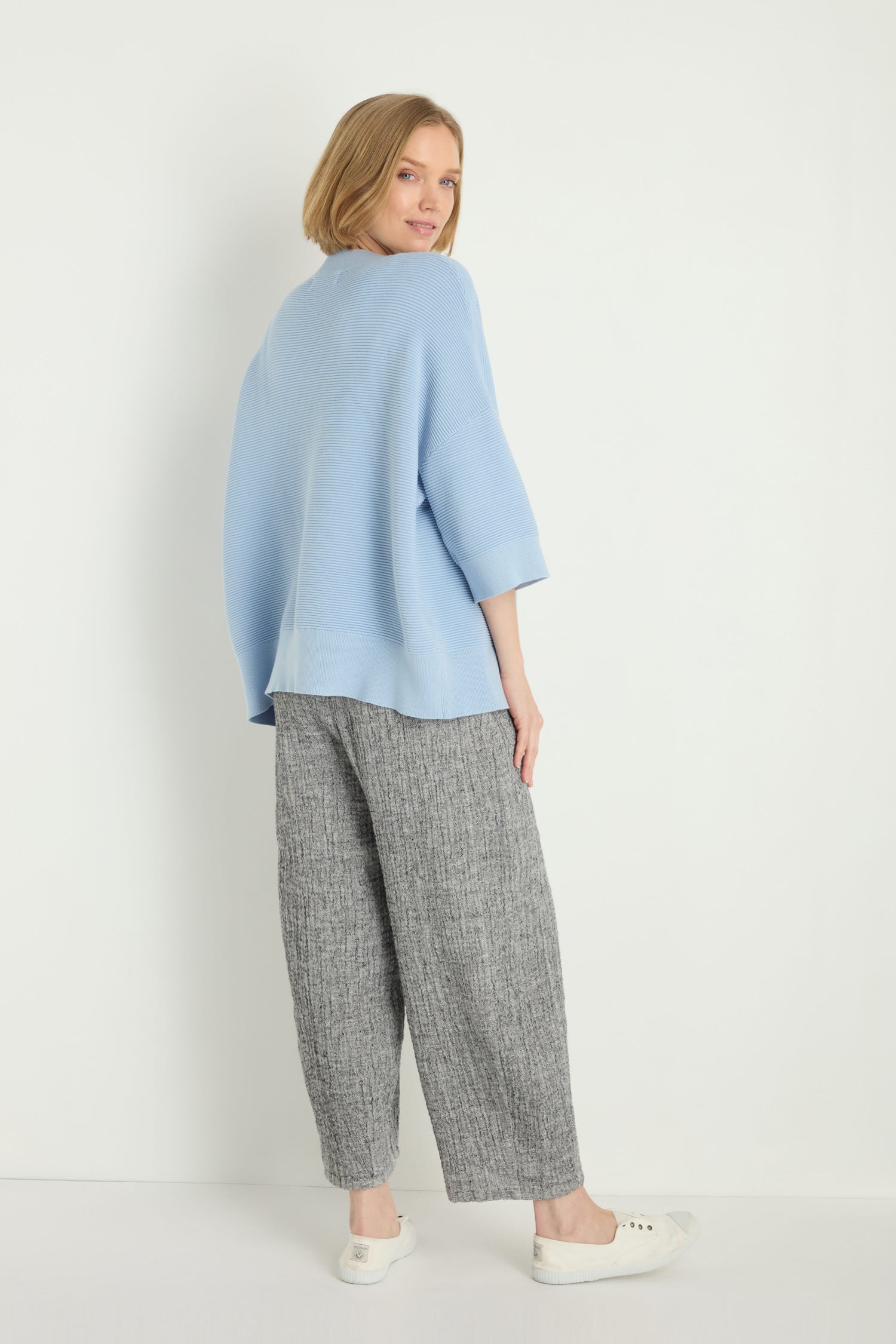A woman poses against a white background wearing a light blue Wide Sleeve Ribbed Top with a boxy silhouette, gray wide-leg pants, and white slip-on shoes. She turns slightly to the side, looking back.