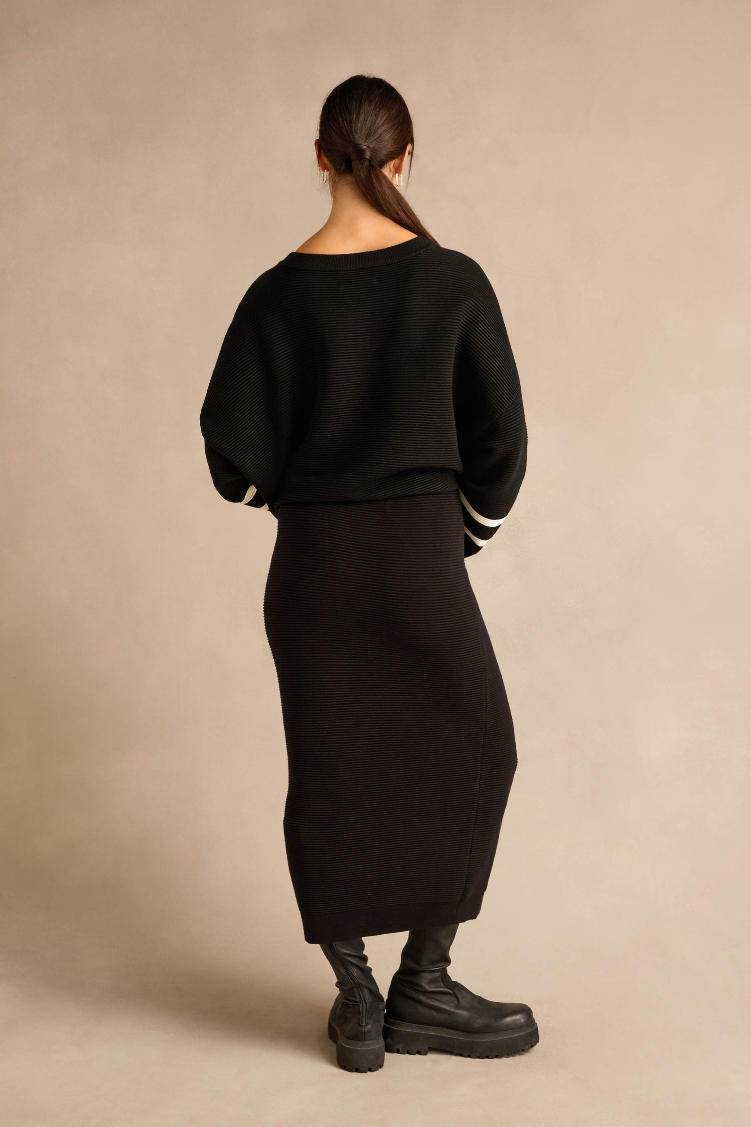 A woman stands facing away, wearing a black sweater, the Ribbed Tube Skirt in a sleek column silhouette that falls to midi length, and black boots, against a plain beige background.