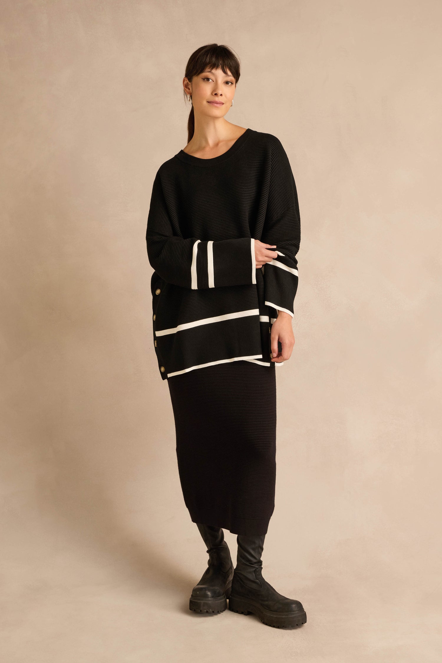 A woman stands against a neutral background wearing the Piping Detail Button Ribbed Top, styled with a black midi skirt and black boots.