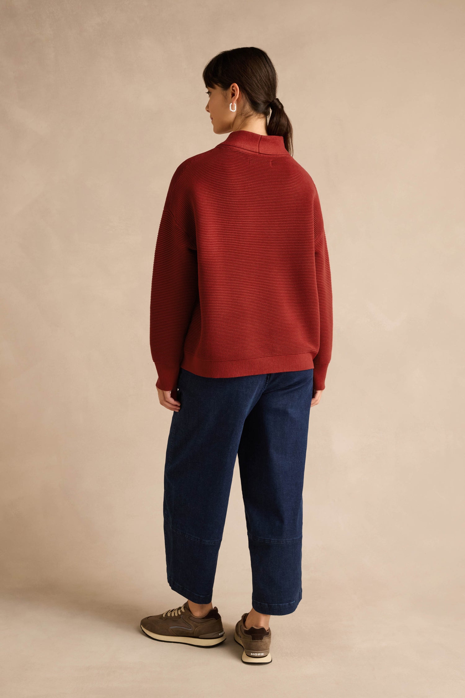 A person stands facing away, wearing the Tie Neck Burgundy Ribbed Knit sweater in a relaxed fit, wide-leg blue jeans, brown sneakers, and round earrings, against a plain beige background.