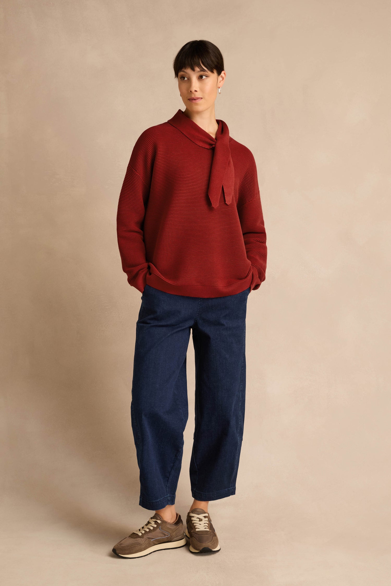 Person standing against a beige backdrop, wearing the Tie Neck Burgundy Ribbed Knit sweater with a relaxed fit, paired with blue loose-fitting pants and brown sneakers.