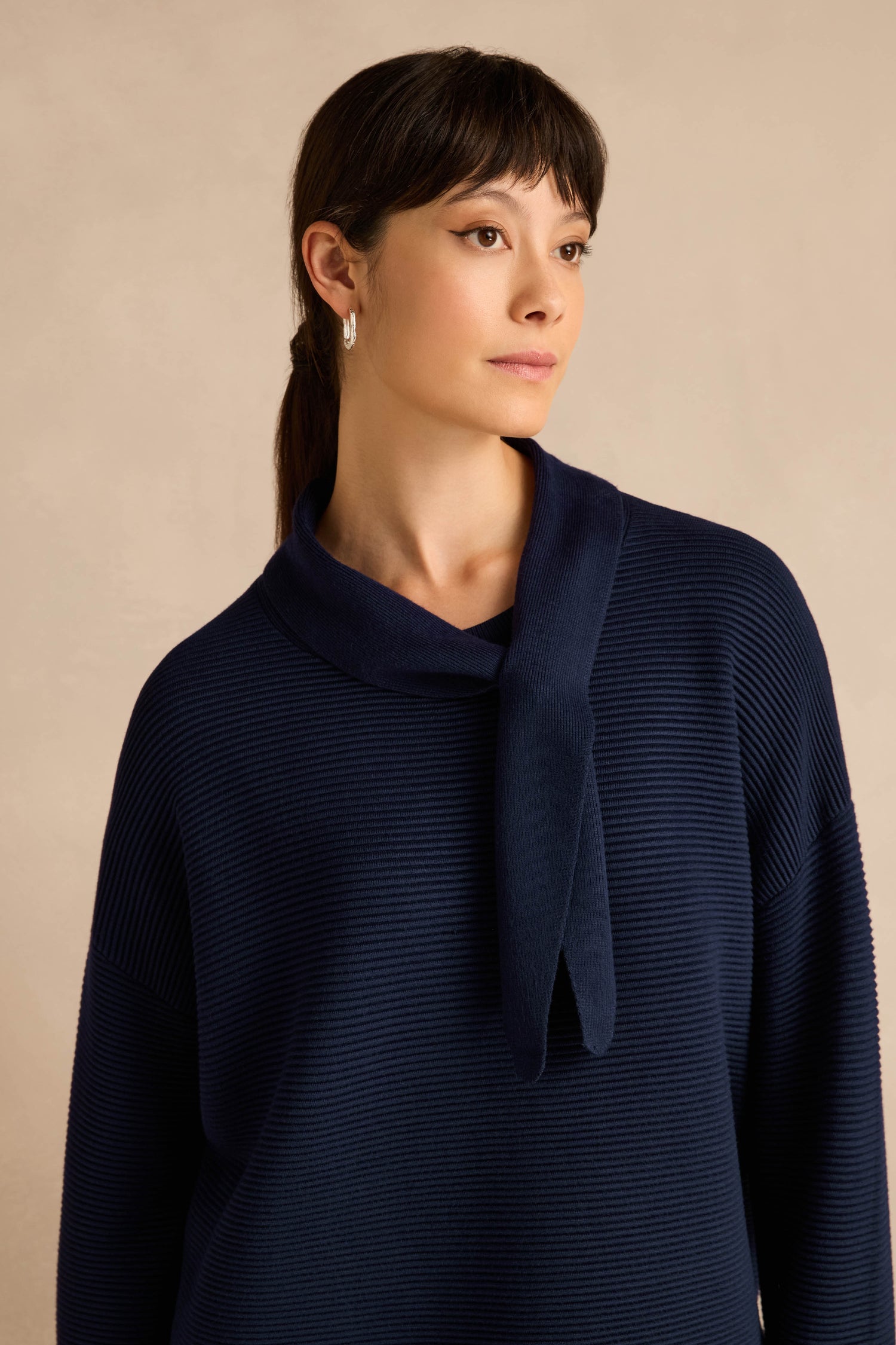 A woman with dark hair in a ponytail wears the Tie Neck Navy Ribbed Knit, standing against a beige background.