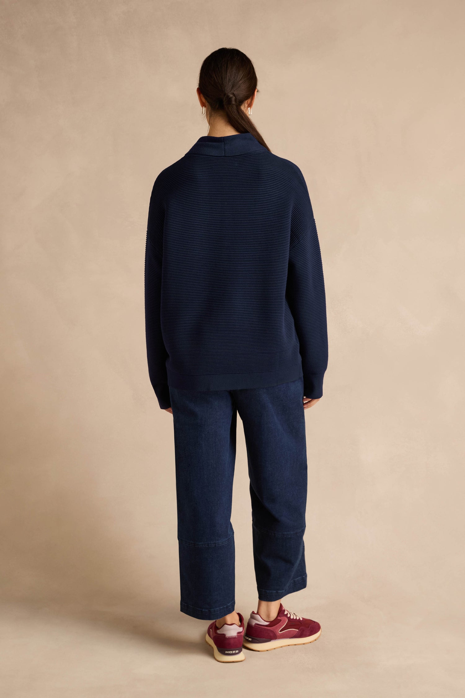 A person stands with their back to the camera against a beige background, wearing the Tie Neck Navy Ribbed Knit in a relaxed fit, paired with wide-leg jeans and maroon sneakers. Their hair is tied back in a low ponytail.