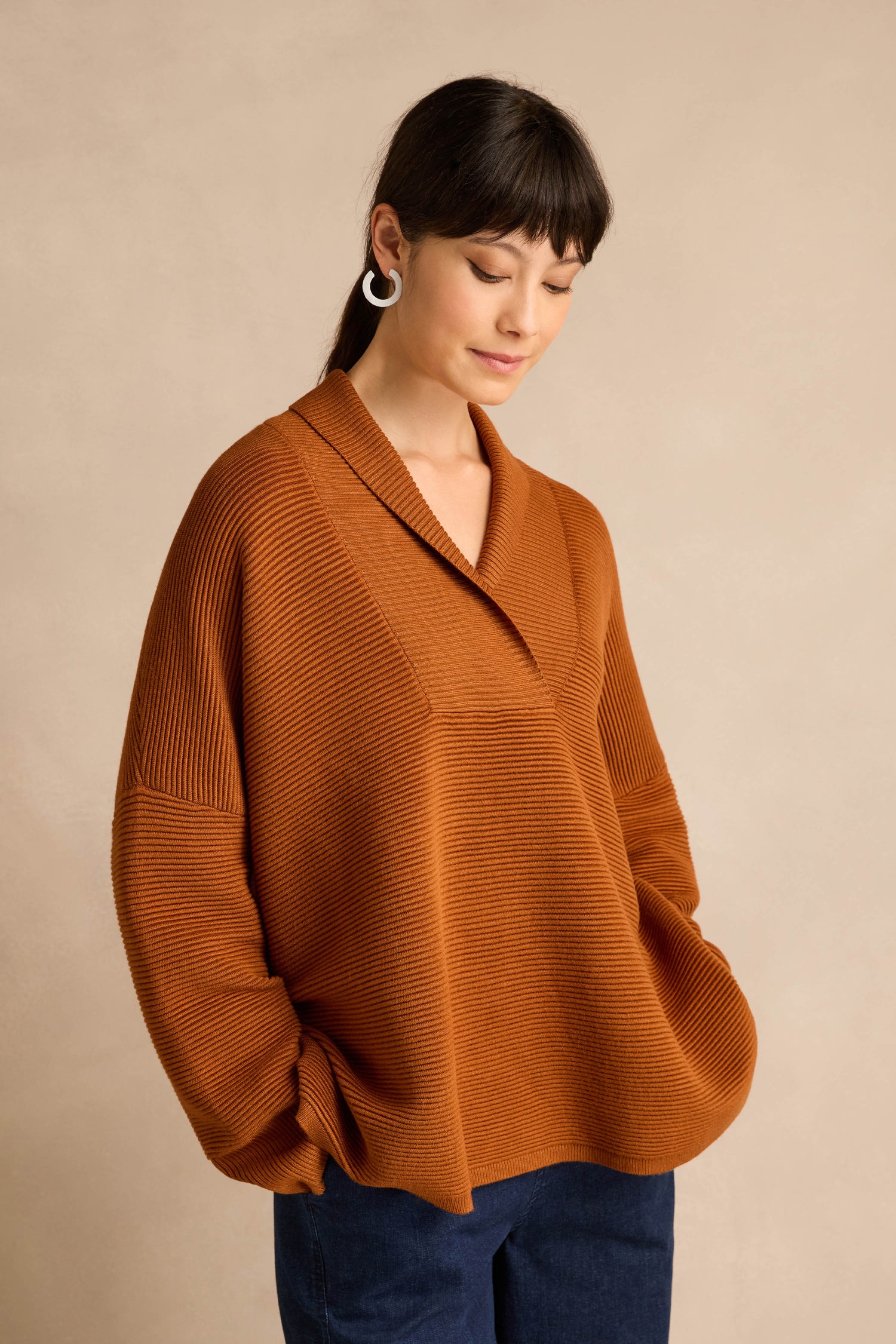 A woman stands against a neutral background with her hands in her pockets, wearing the Shawl Collar Copper Ribbed Knit—a loose, long-sleeved sweater featuring a ribbed texture and rich copper hue.