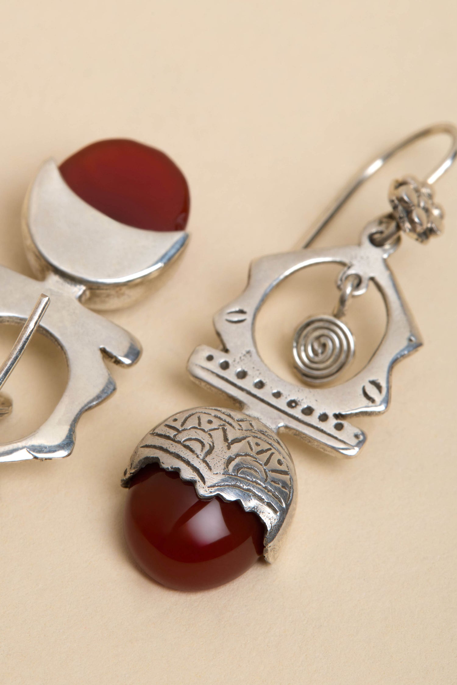 Close-up of Carnelian Drop Earrings and a brooch with red stones on beige. The jewelry features engraved patterns and geometric designs.