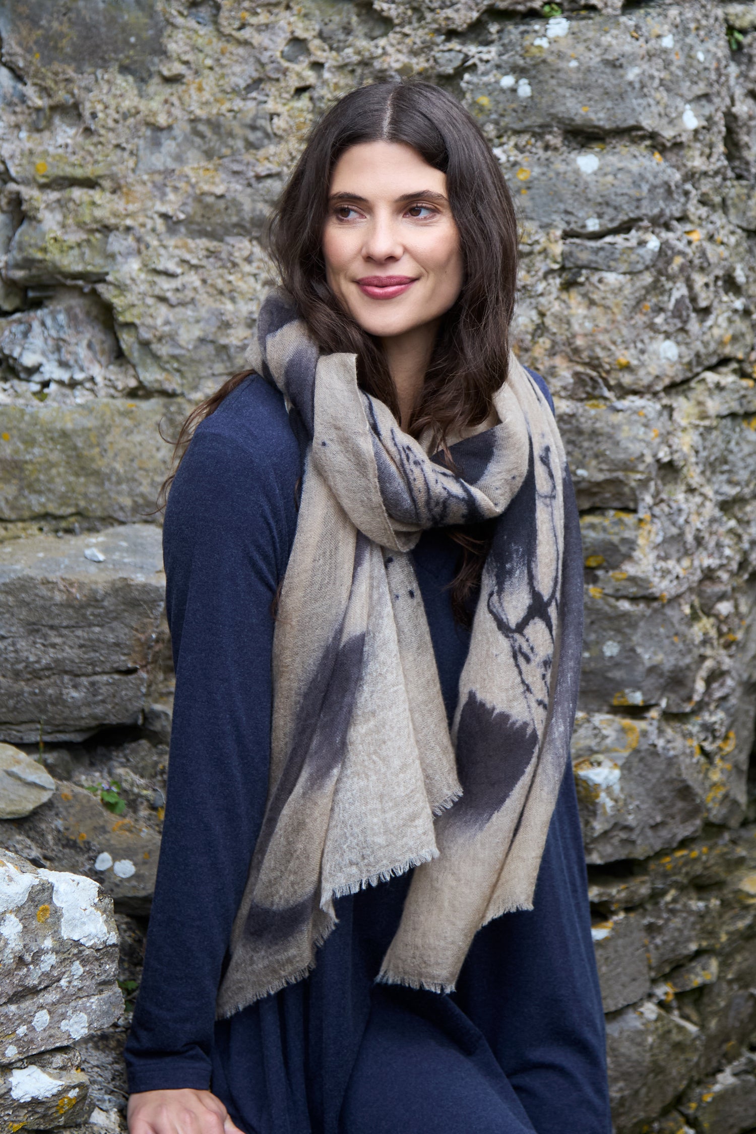A woman with long brown hair in a dark blue dress wears the Painted Brushstroke Cashmere Scarf, featuring an abstract pattern, as she sits against a stone wall and gazes slightly to the side.