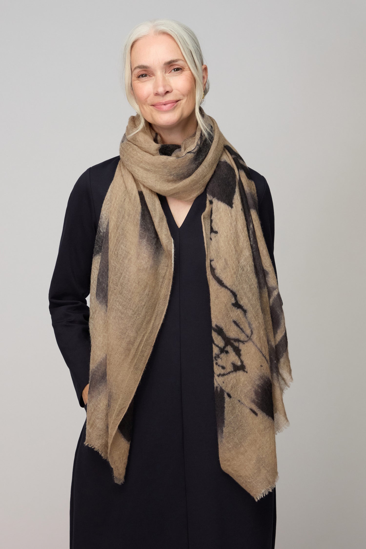 A woman with long gray hair in a low ponytail stands against a plain background, wearing the Painted Brushstroke Cashmere Scarf—beige with black brushstroke design—draped over a black long-sleeve dress, her hands in her pockets.