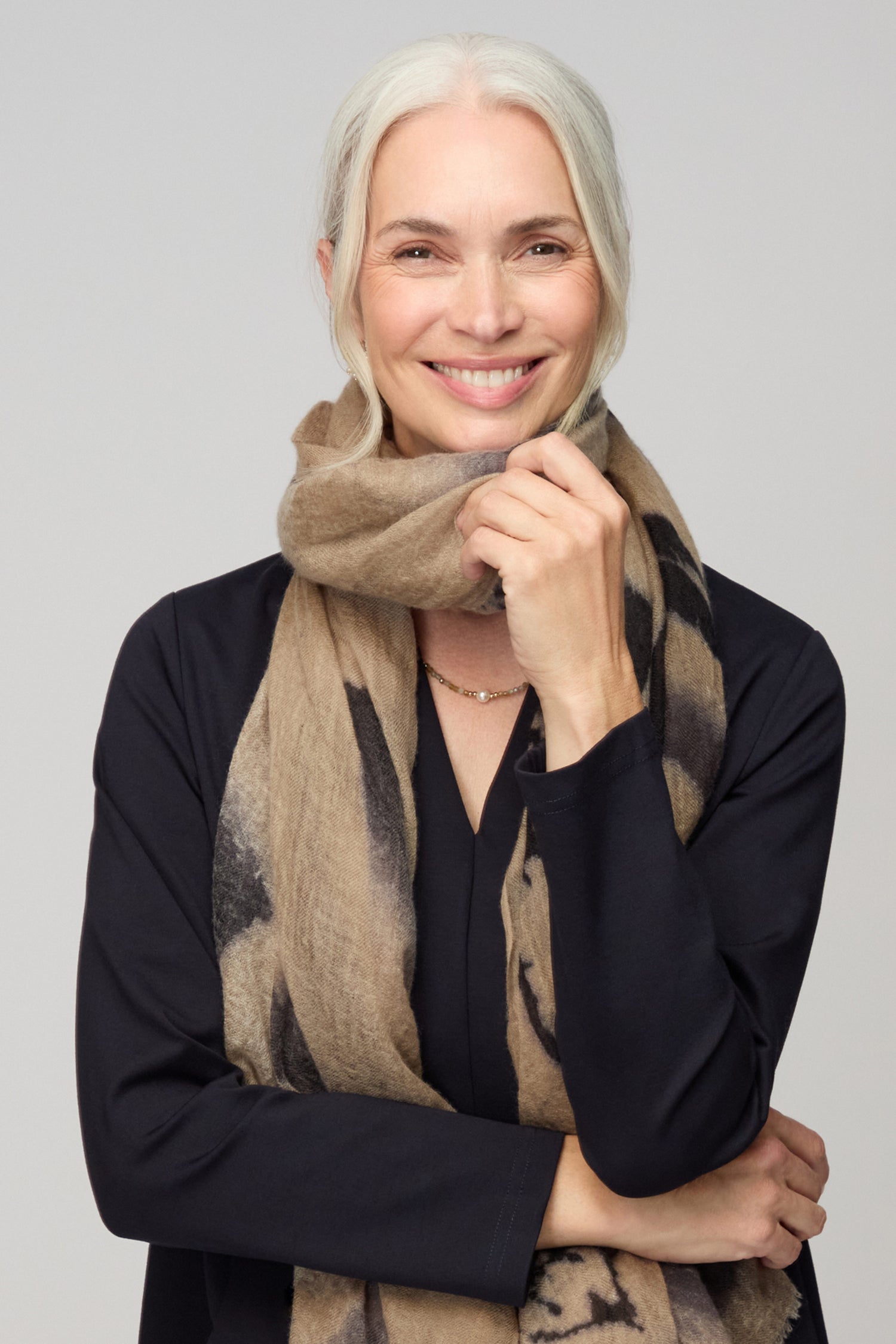 Smiling older woman with white hair, wearing a black top and the Painted Brushstroke Cashmere Scarf, poses with one hand touching her scarf against a plain background.