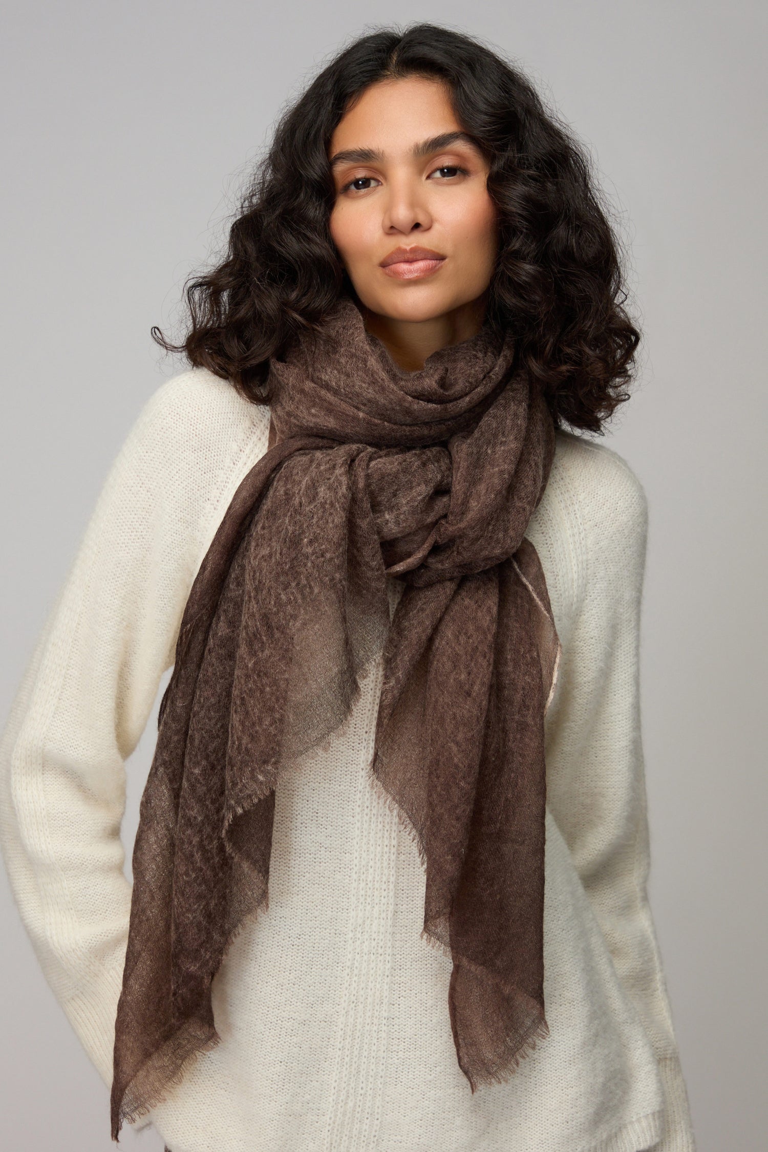A woman with wavy dark hair wears a white sweater and the Soft Espresso Cashmere Scarf, standing against a plain light gray background.