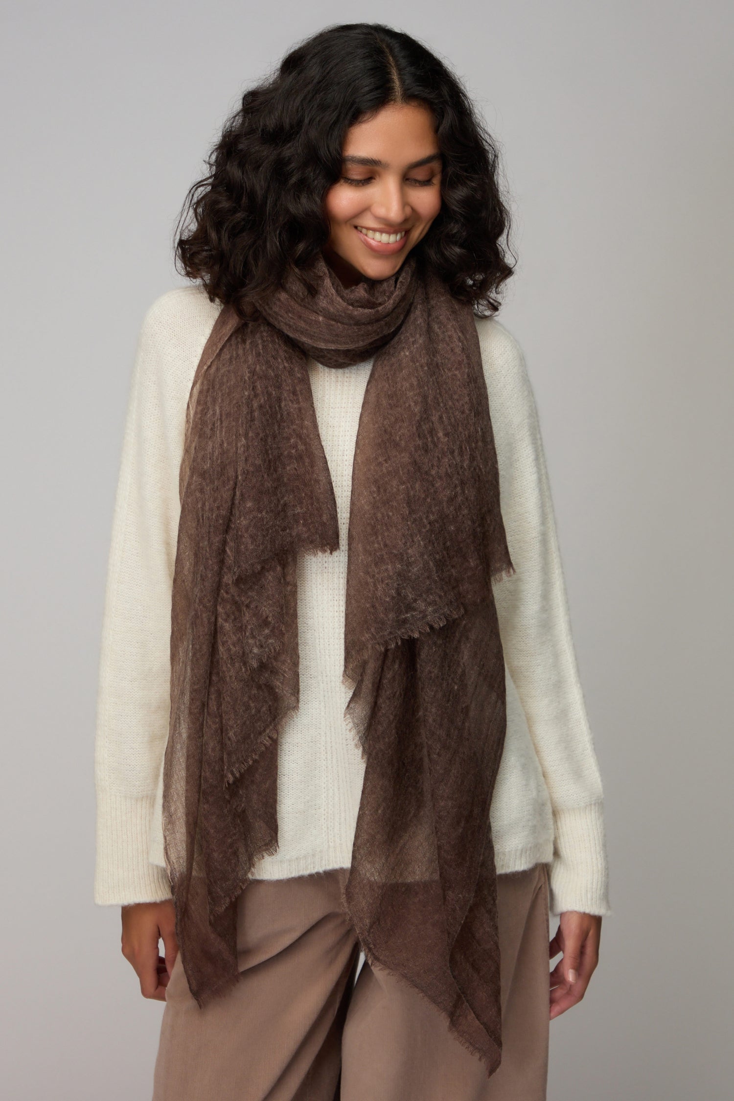 A woman with curly dark hair wears a white sweater, taupe pants, and the Soft Espresso Cashmere Scarf, standing against a plain background and smiling while looking down.