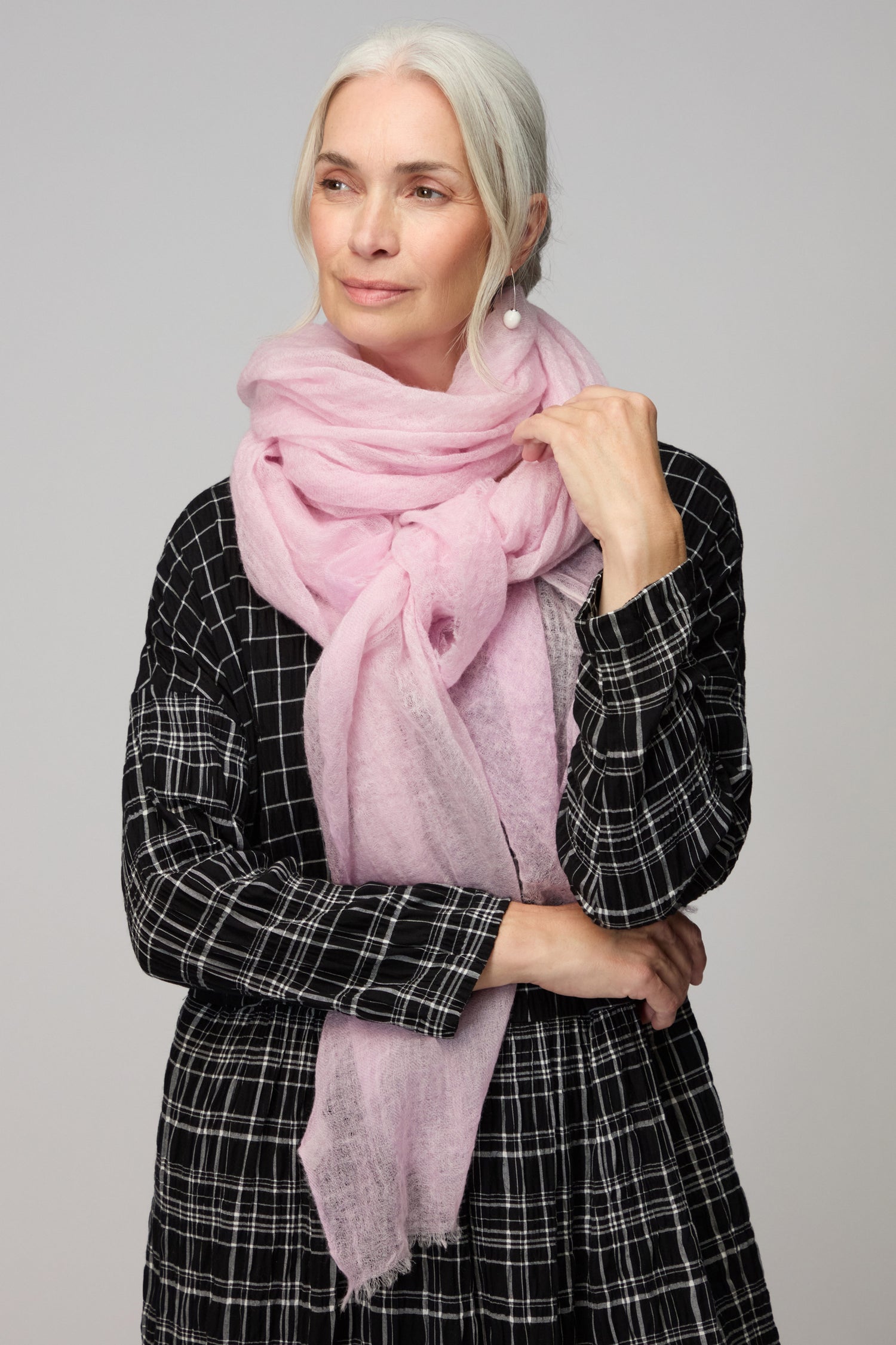 A woman with white hair wears a black plaid dress and a Soft Blush Gradient Cashmere Scarf, standing against a light background to showcase this perfect lightweight accessory for any outfit.