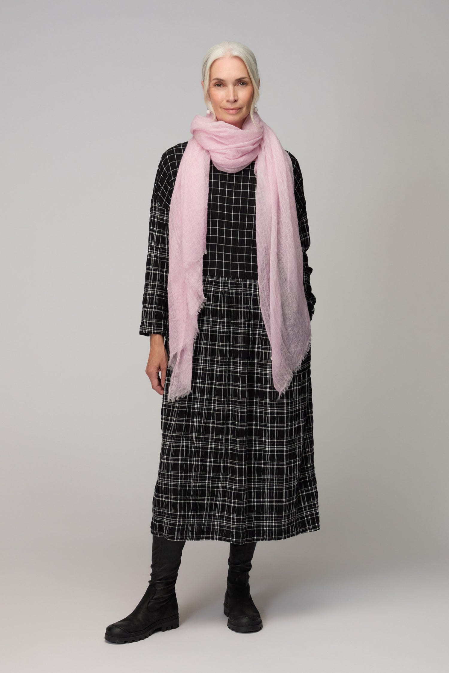 A woman with gray hair faces forward, wearing a black checkered dress, black boots, and the Soft Blush Gradient Cashmere Scarf—a lightweight accessory that adds a gentle touch to her ensemble.