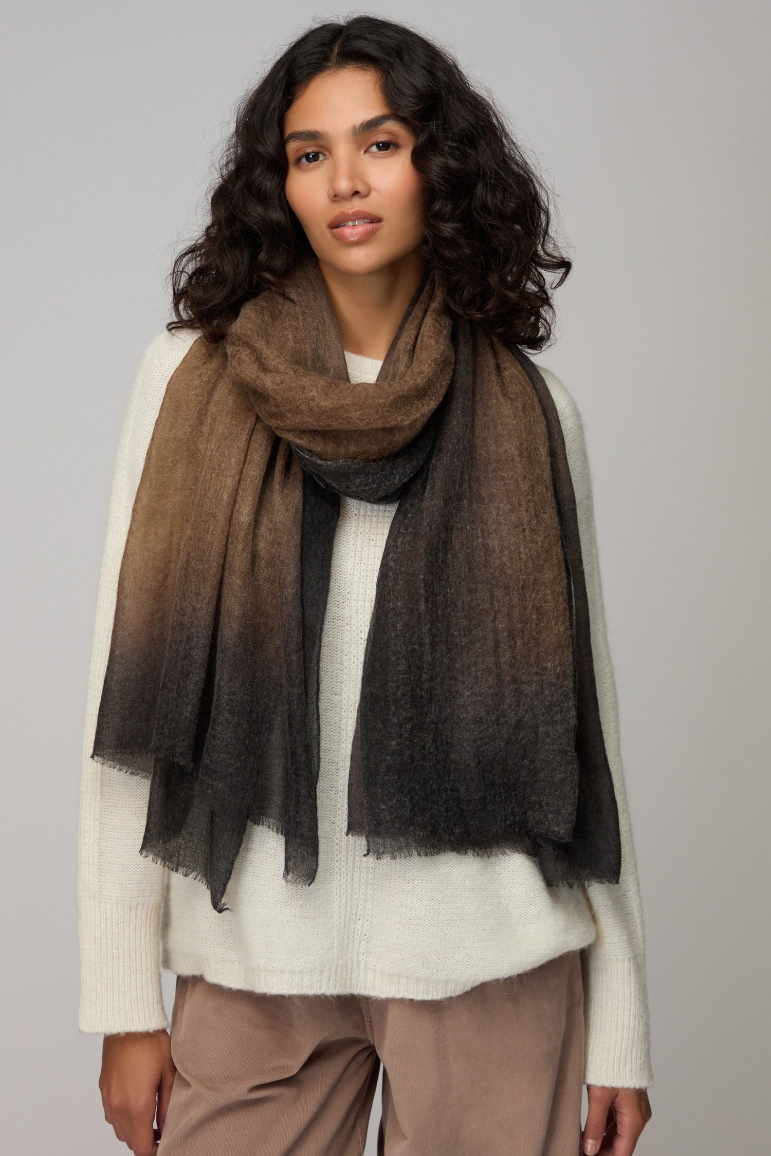 A woman in a cream sweater and light brown pants stands against a plain light background, accessorized with the Espresso Gradient Cashmere Scarf—a luxurious brown-to-black gradient piece.