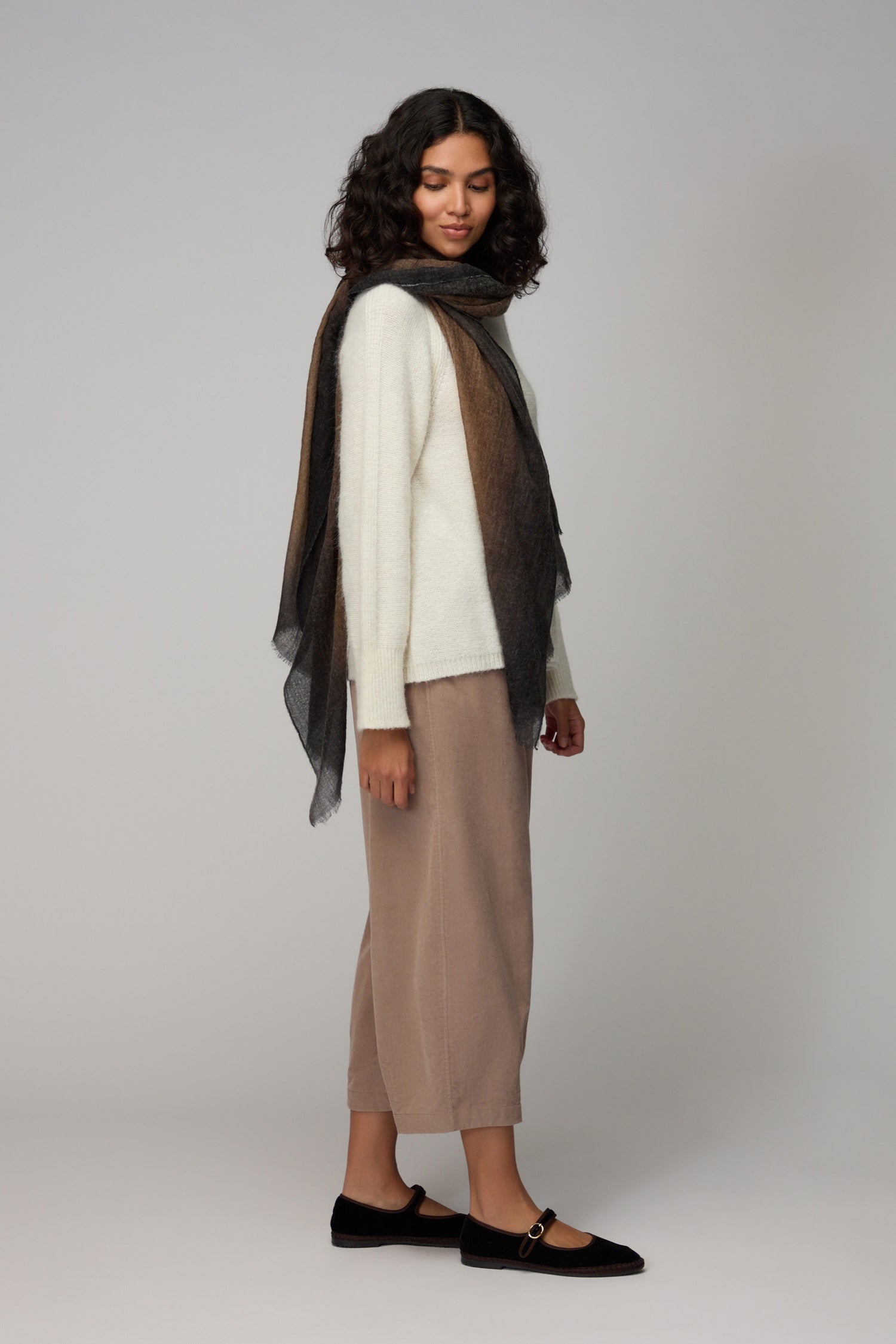 Woman standing in profile against a plain light background, wearing a white sweater, taupe pants, black flats, and the Espresso Gradient Cashmere Scarf—a luxury accessory.