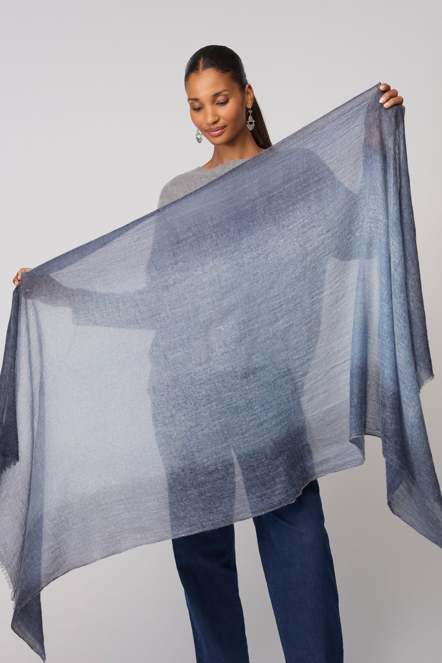 A woman in a gray top and blue pants showcases the Night Sky Gradient Cashmere Scarf, featuring lightweight fabric in shades of gray and navy, against a plain background.