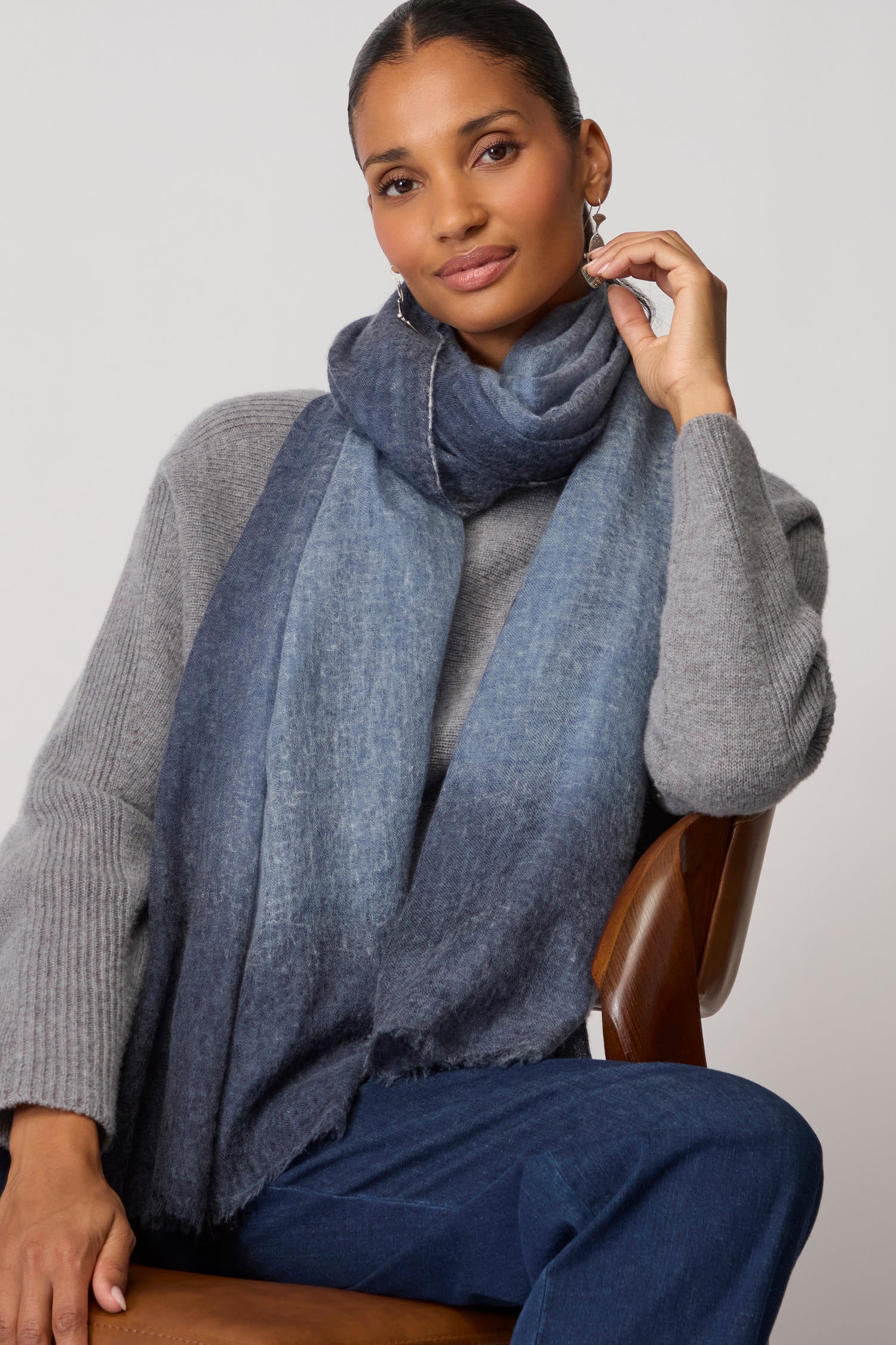 A woman seated on a chair wears a grey sweater, blue jeans, and the Night Sky Gradient Cashmere Scarf, with one hand touching her earring.
