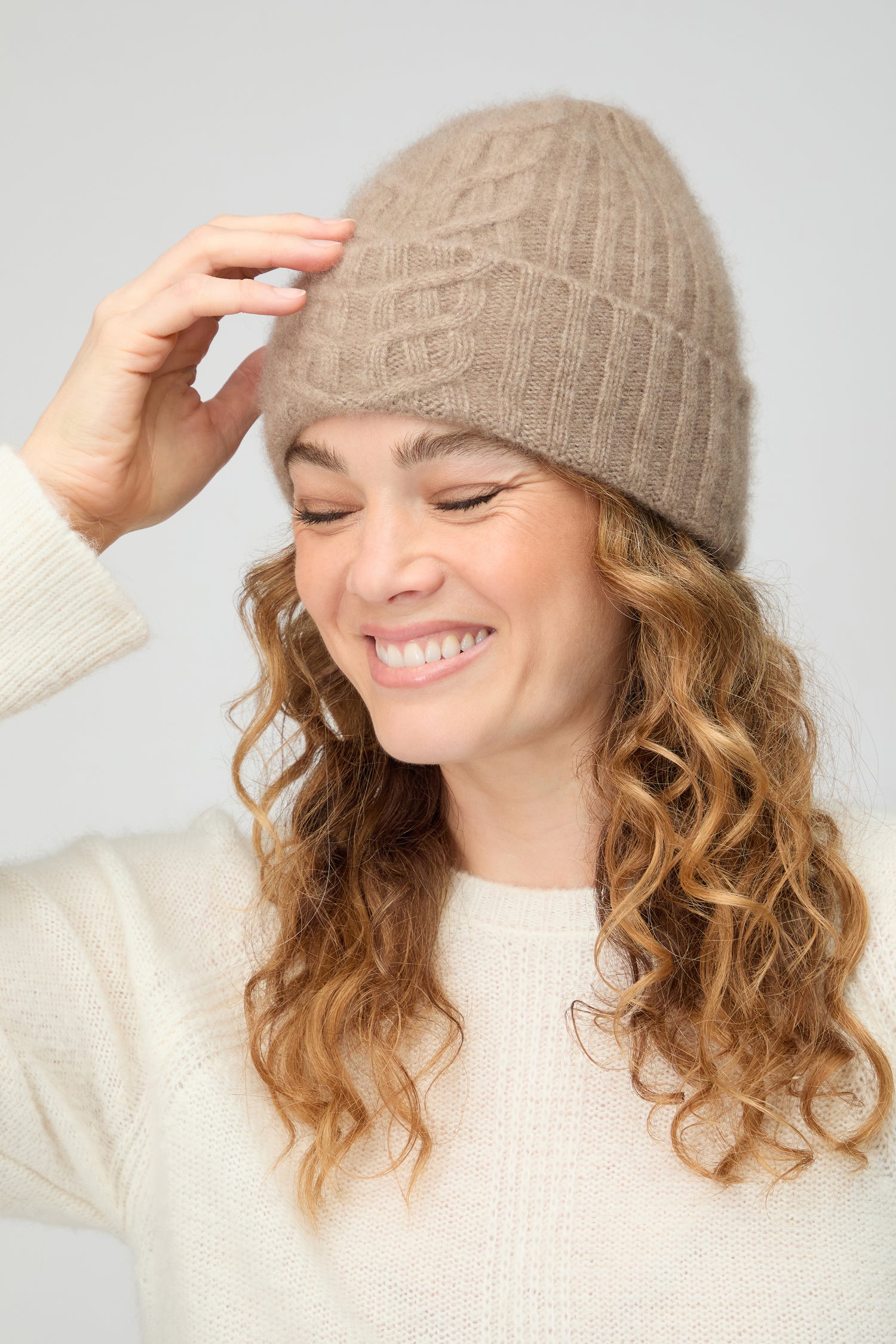 A woman with curly hair, wearing the Warm Sands Ribbed Cashmere Hat and a cream sweater, smiles with her eyes closed while touching the side of her winter accessory.