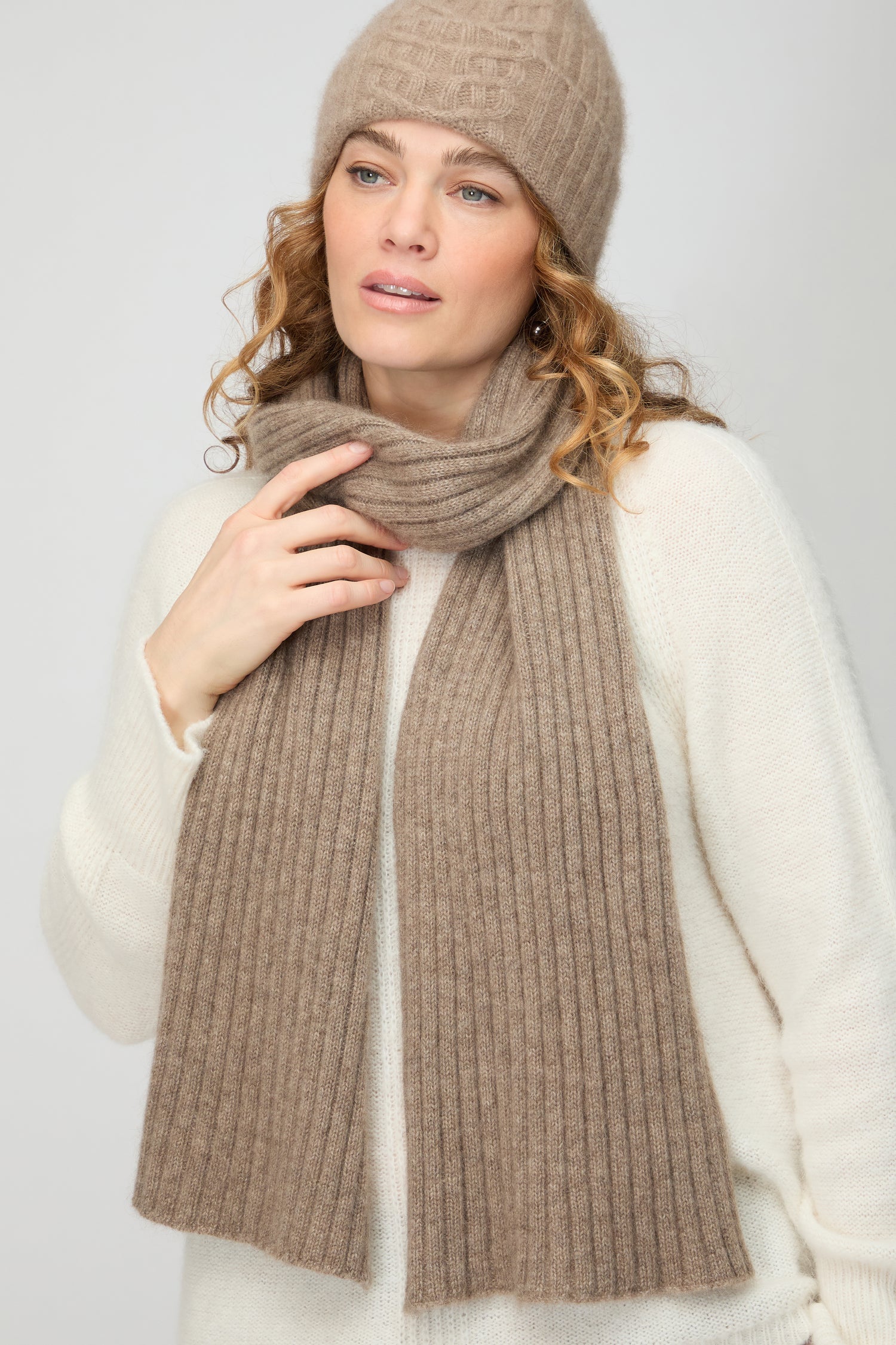 A woman wears the Warm Sands Ribbed Cashmere Scarf and a beige knit hat over a cream sweater, gazing to the side against a plain background—an effortlessly chic addition to your winter wardrobe.