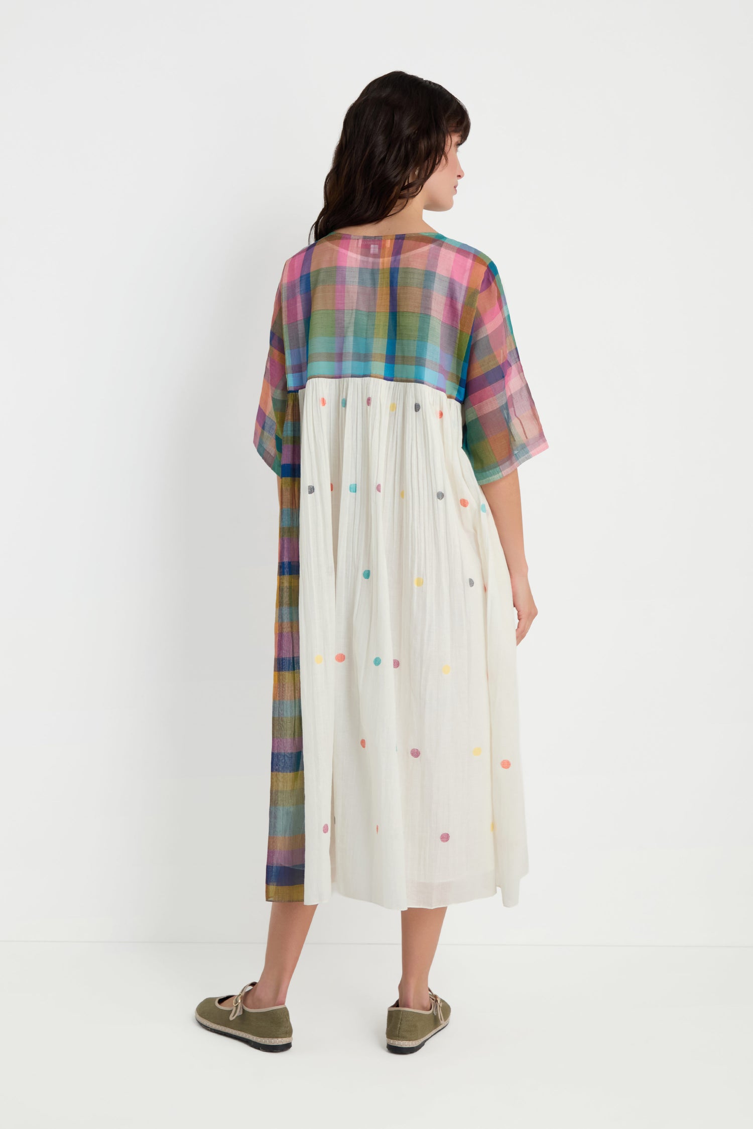 A woman stands with her back to the camera wearing the Mondrian Jamdani Dress, a midi dress crafted from jamdani cotton with a colorful plaid top and white skirt detailed with multicolored dots.