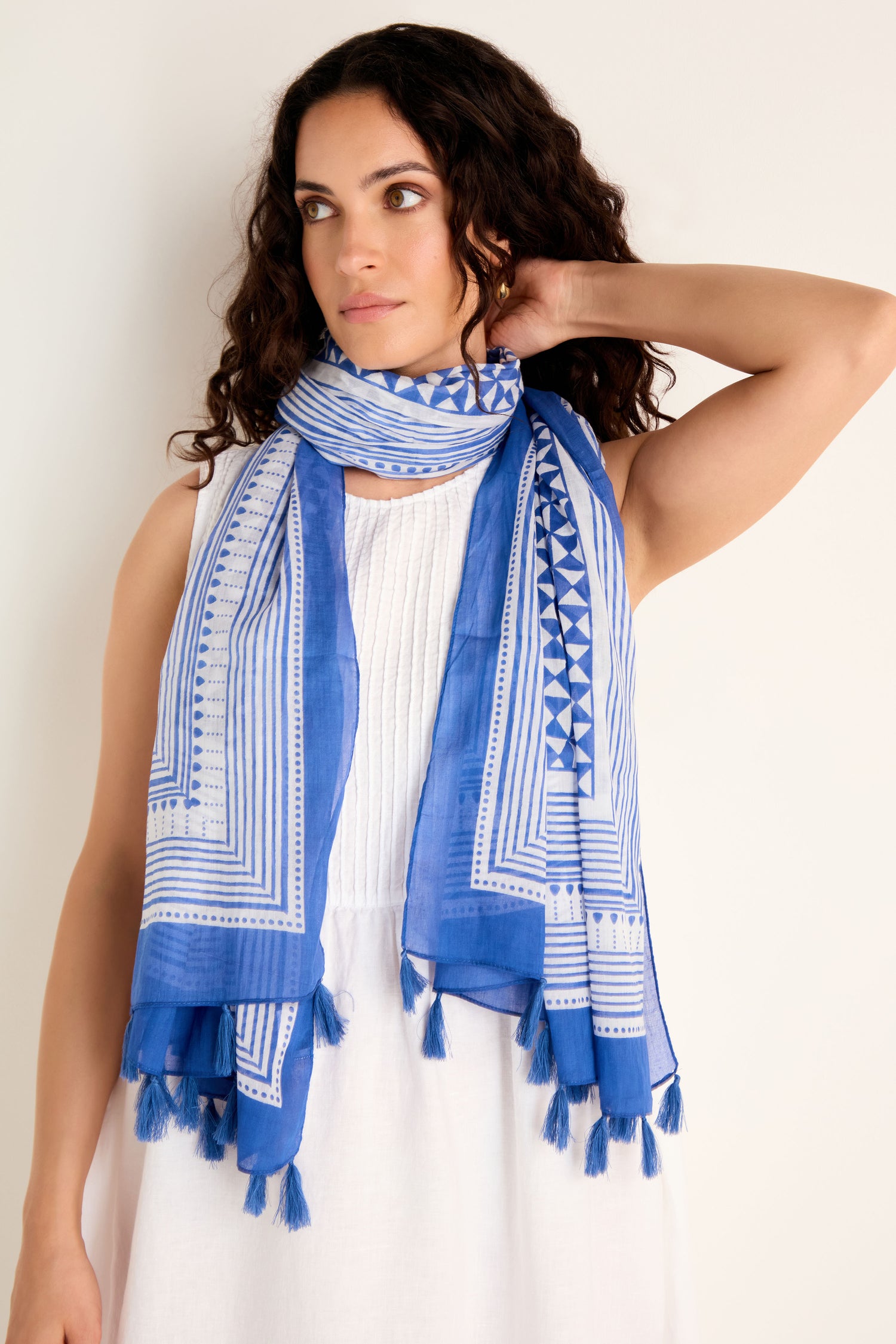 A woman stands against a plain light background, wearing the Cobalt Tassel Cotton Scarf featuring a blue and white geometric print and tassel edging, draped over a sleeveless white dress.
