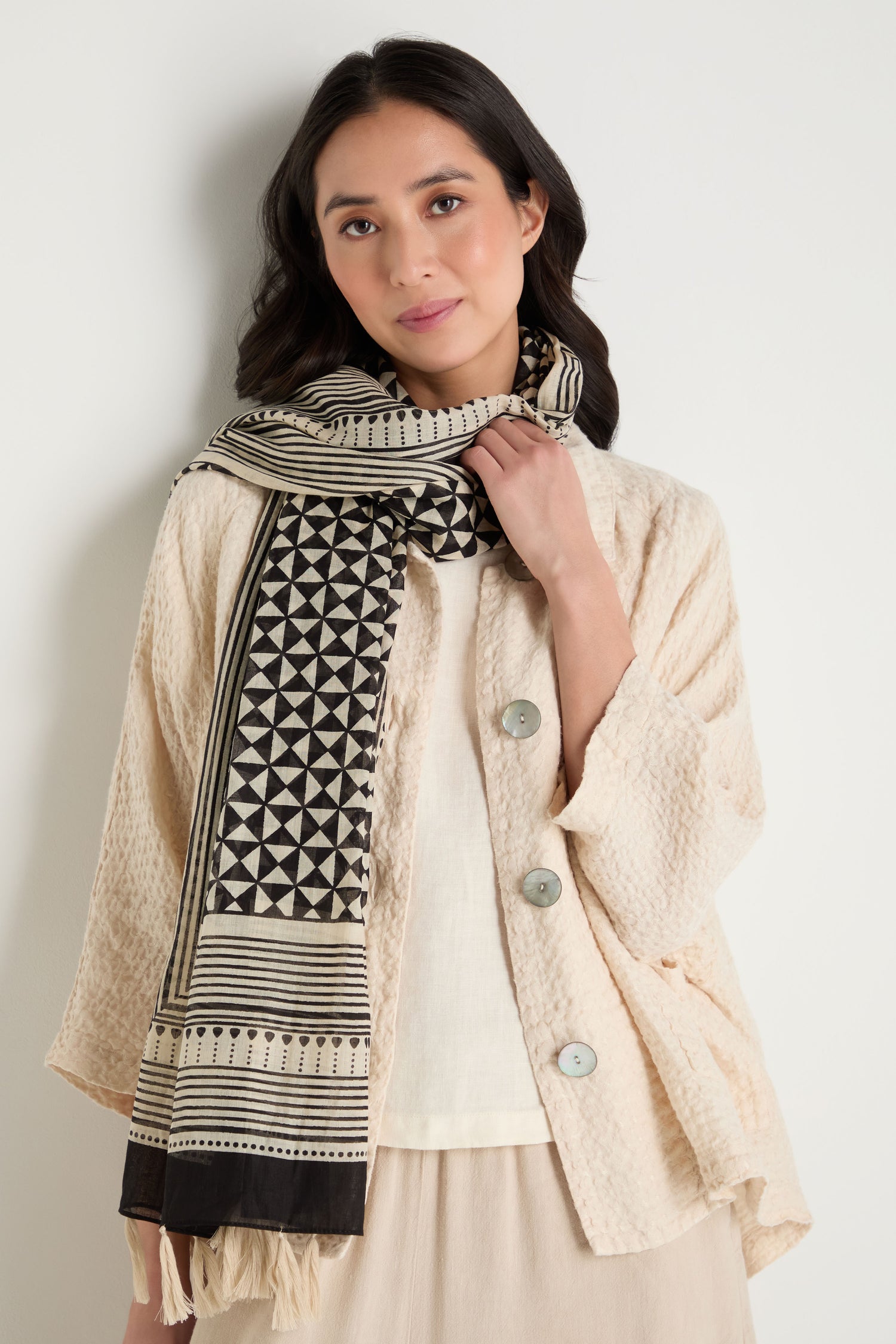 A woman with long dark hair wears a cream textured jacket over a white top and a Cotton Tassel Scarf, standing against a plain white background.