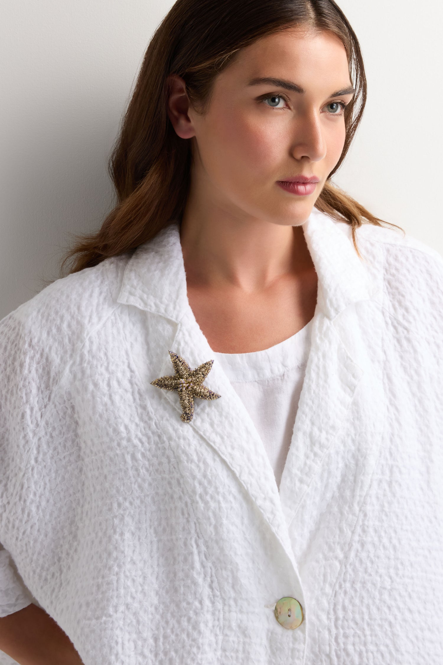 A woman stands against a plain white background, wearing a textured white jacket adorned on the lapel with the Hand Beaded Starfish Brooch, featuring large crystal beads.