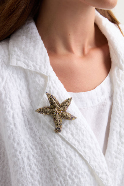 A person wears a textured white jacket and top, with the Hand Beaded Starfish Brooch—adorned with shimmering crystal beads—pinned to the lapel.