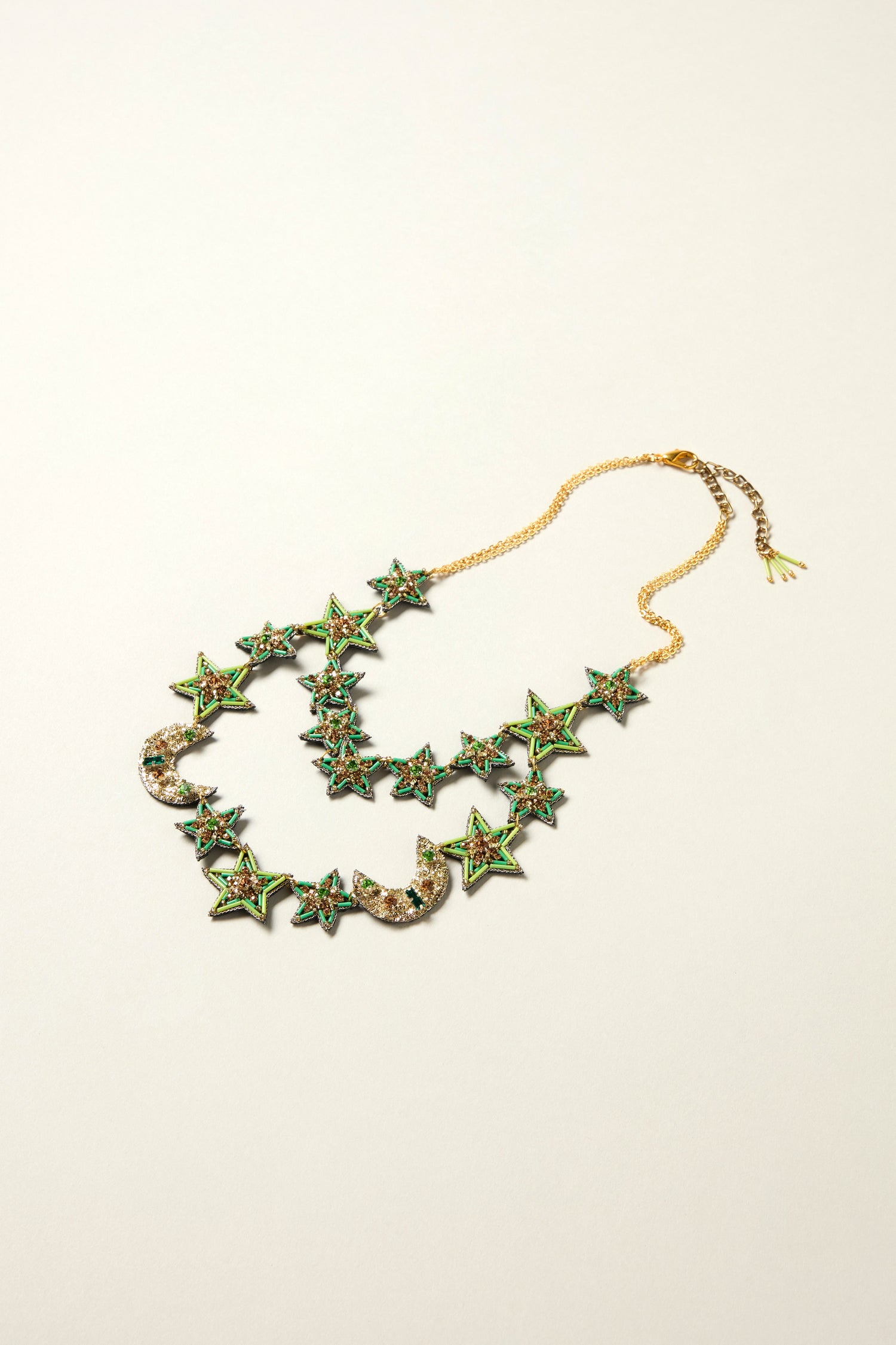 The Hand Beaded Celestial Necklace features double strands with green star and crescent moon pendants adorned with colorful stones, shown on a neutral background—a beautifully handcrafted jewelry piece.