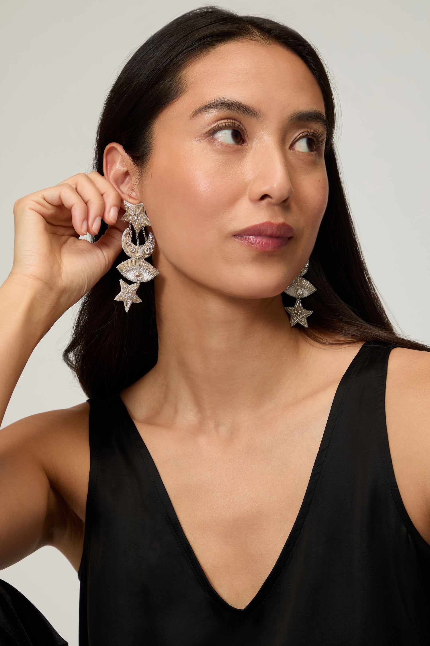 A woman with long dark hair in a sleeveless black top models Psquuare Fashion Jewellery’s Hand Beaded Celestial Silver Earrings, holding one earring in her hand.