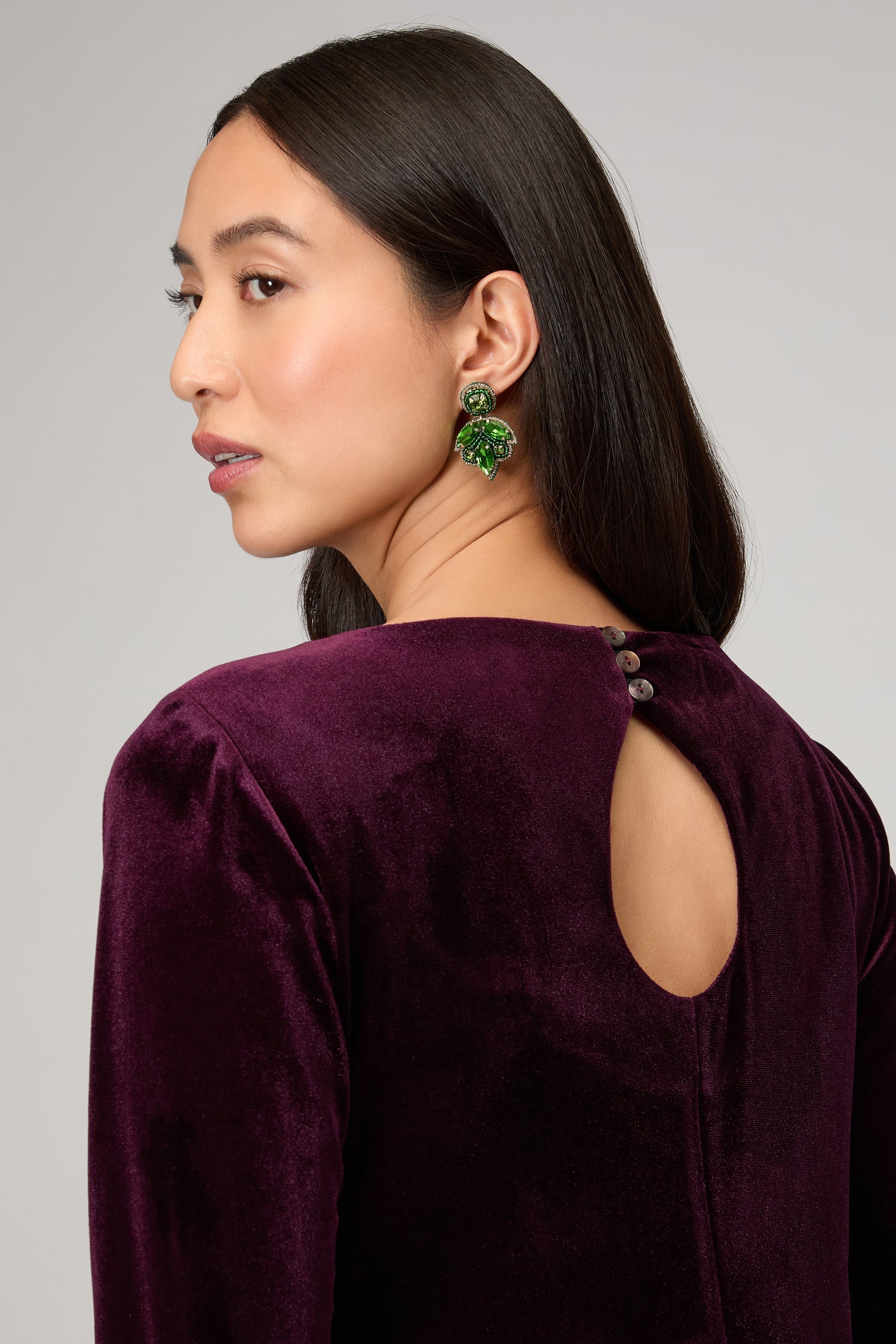 A woman with long dark hair wears a velvet purple dress and Hand Beaded Flower Earrings—lightweight statement jewelry in vivid green—while looking over her shoulder.