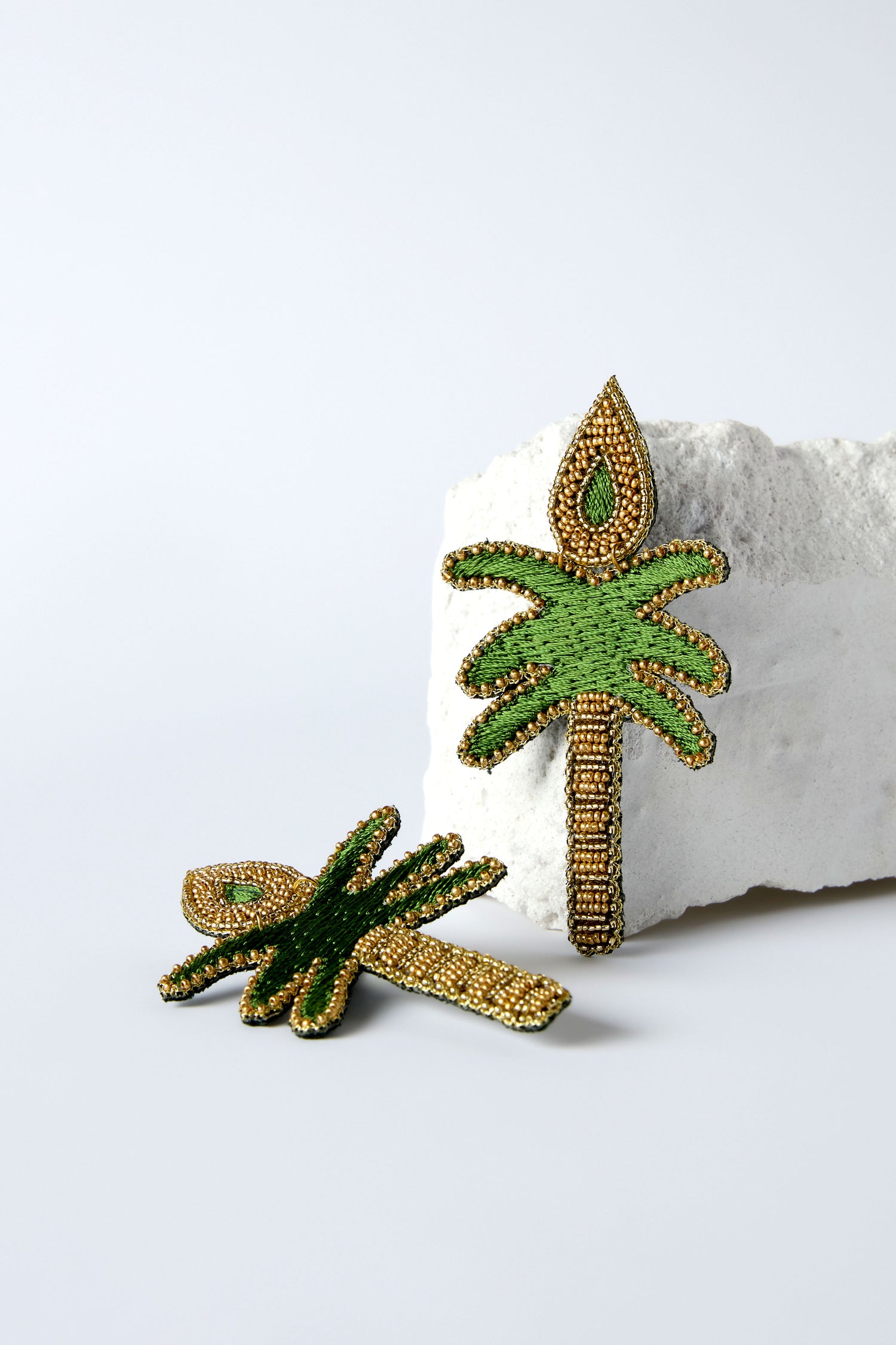 Hand Embroidered Palm Earrings in green and gold beading are shown—one flat, one propped on a white stone—ideal for lovers of unique, hand-beaded palm earrings.