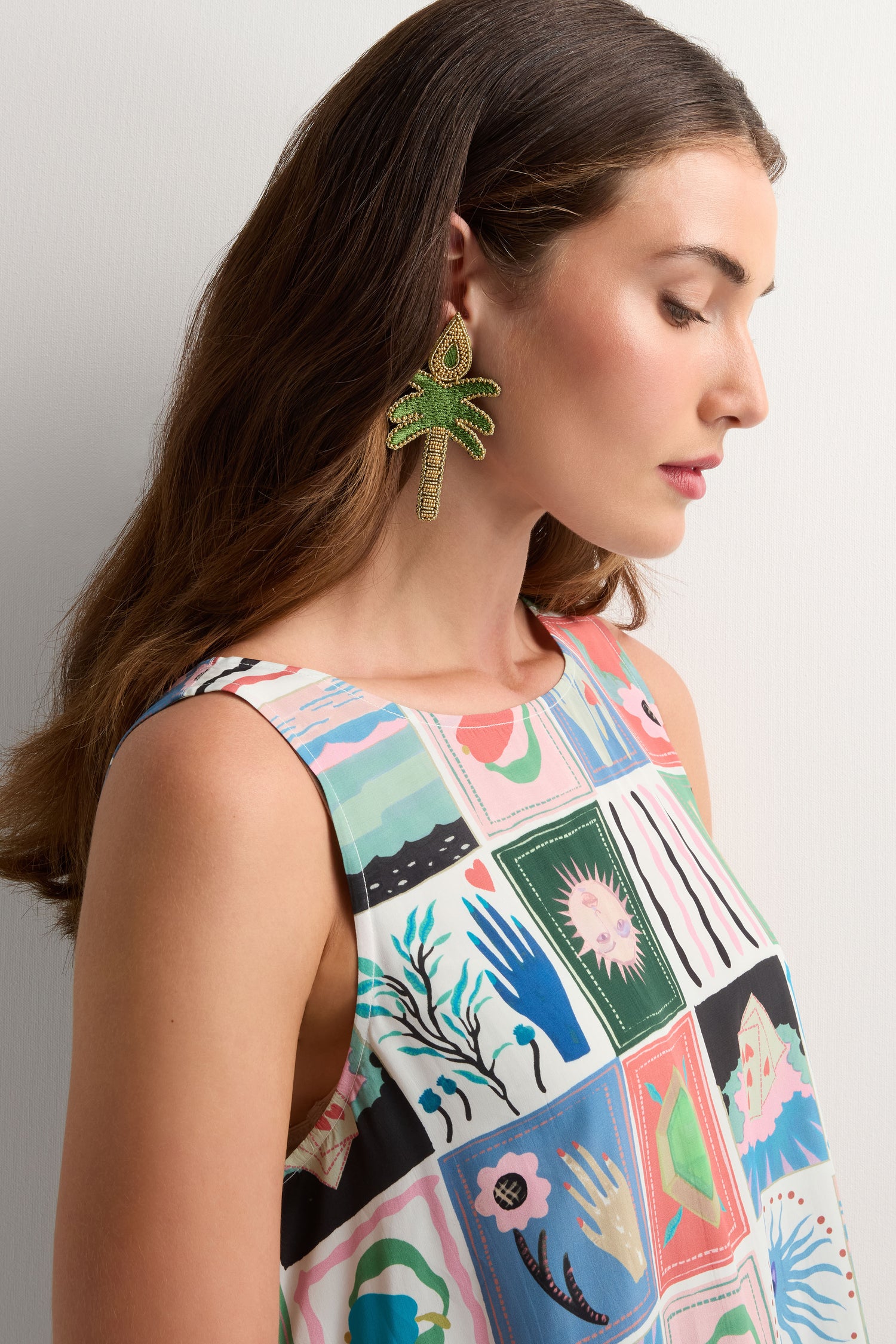 Woman in a sleeveless, patterned dress with abstract designs, shown in profile against a plain background, wearing large Hand Embroidered Palm Earrings shaped like green leaves.