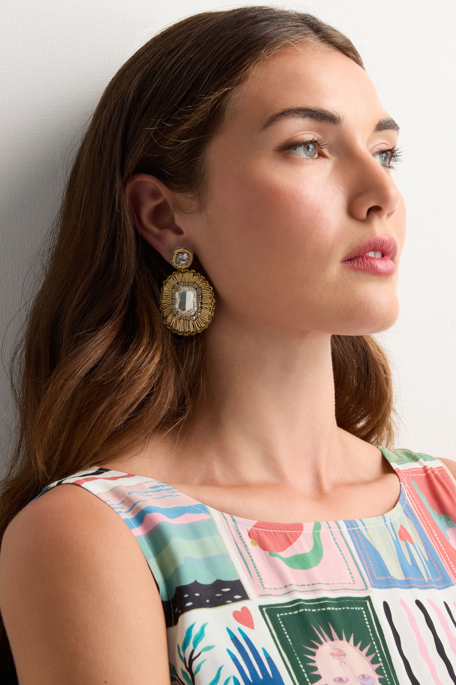 A woman with long brown hair wears a patterned sleeveless top and Hand Embroidered Ornate Earrings featuring crystal beads, gazing slightly to the side against a plain background.