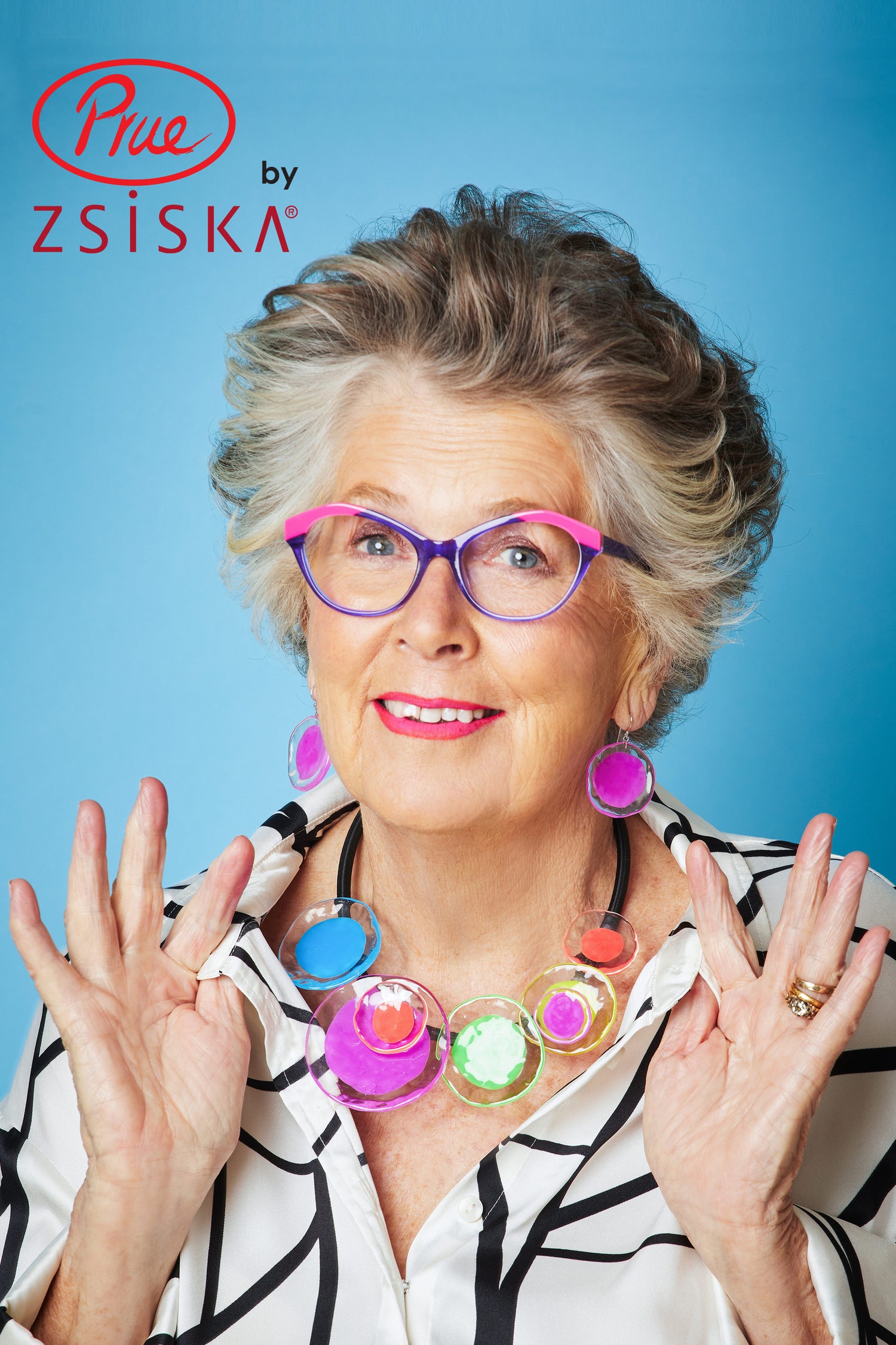 An elderly woman with gray hair, colorful glasses, and a necklace raises her hands in joy to highlight her Flora Earrings. The backdrop is blue with a logo text in the top left.
