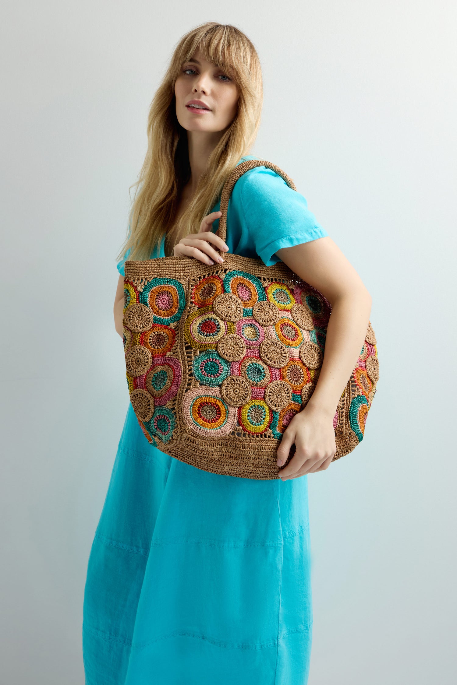A person in a bright blue dress holds the Handwoven Circles Raffia Bag, featuring large, colorful circular patterns, exuding bohemian charm.