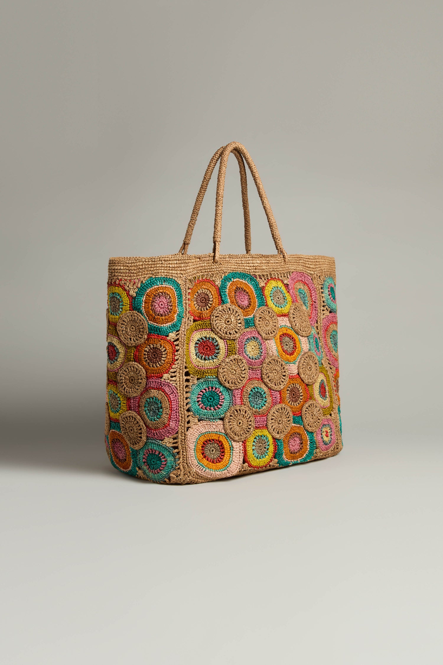 The Handwoven Circles Raffia Bag is a large woven tote showcasing round, multicolored crochet patterns that exude bohemian charm. With two sturdy handles, this raffia bag elegantly rests against a neutral background.