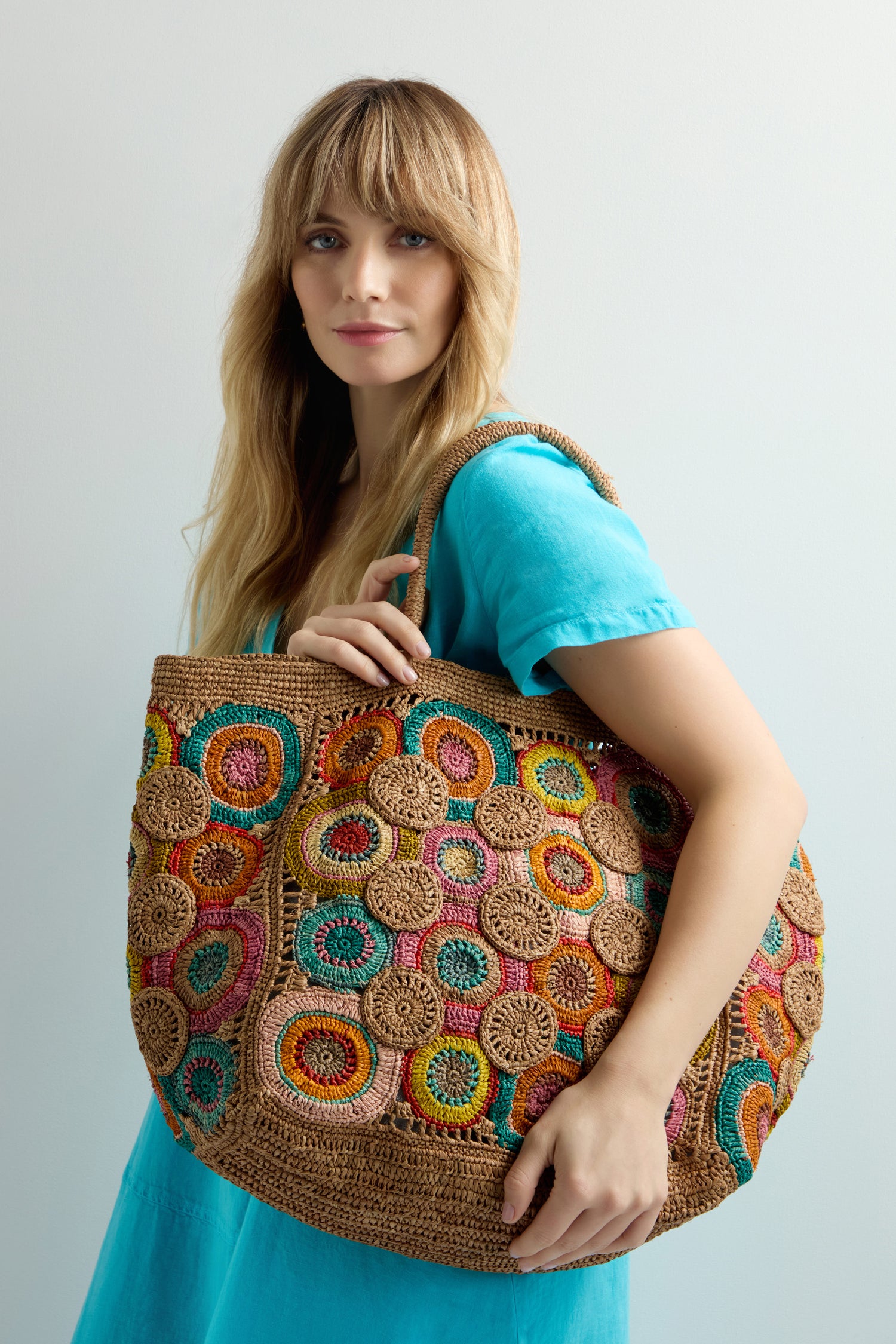 A person with long hair in a turquoise dress holds a large, multicolored Handwoven Circles Raffia Bag, exuding bohemian charm against a plain background.