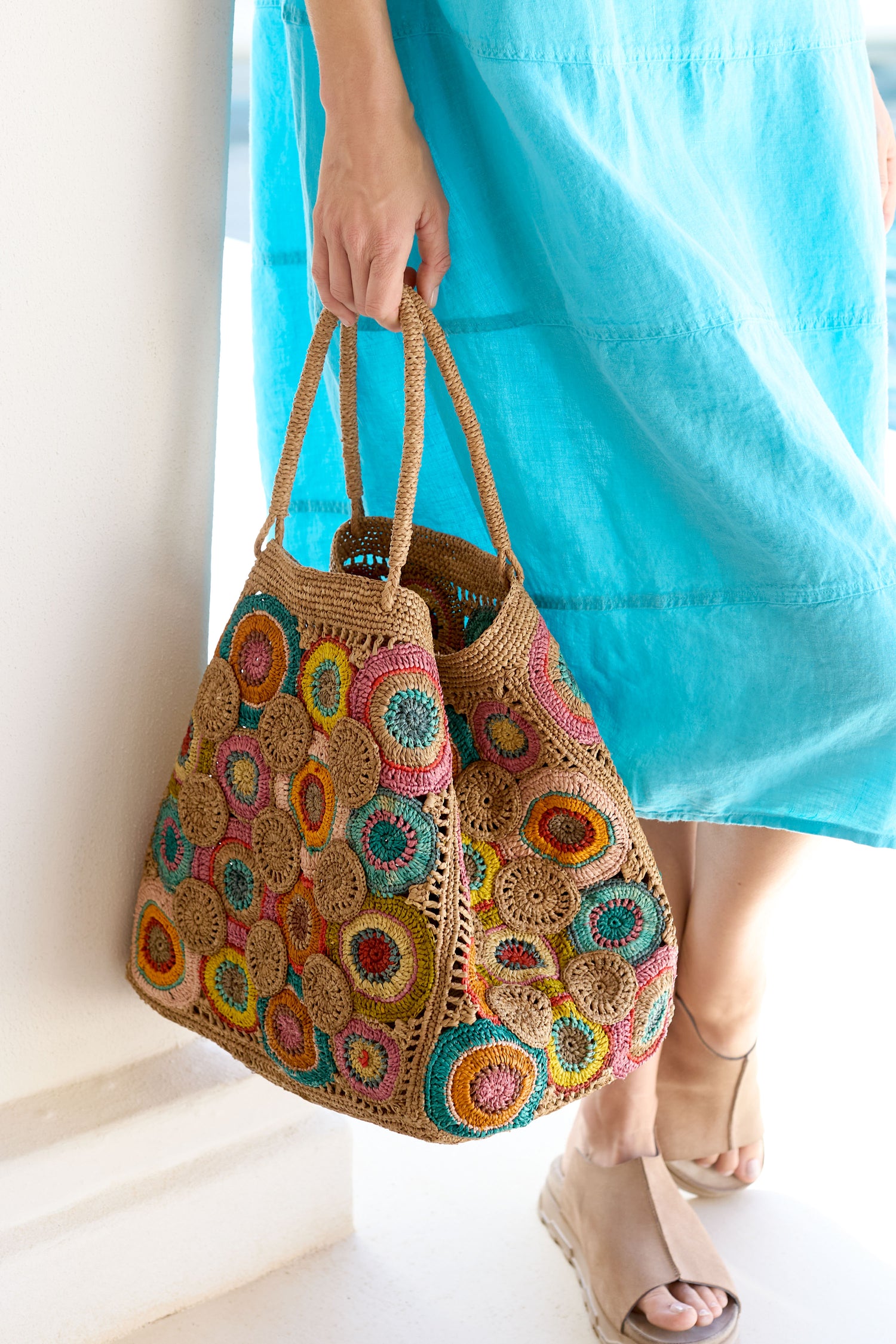 A person is holding a Handwoven Circles Raffia Bag, wearing a light blue dress paired with beige wedge sandals.
