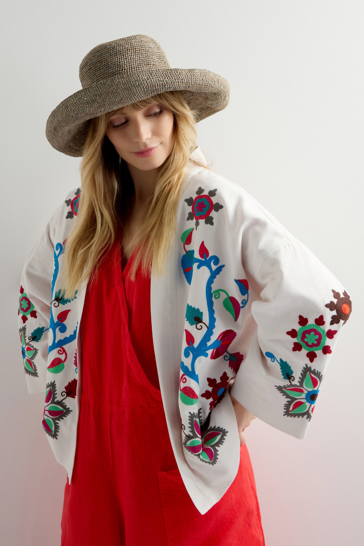 Wearing a Handwoven Wide Brim Raffia Hat for sun protection, a person pairs it with a vibrant red outfit and a white jacket featuring colorful floral embroidery, standing against a plain backdrop.