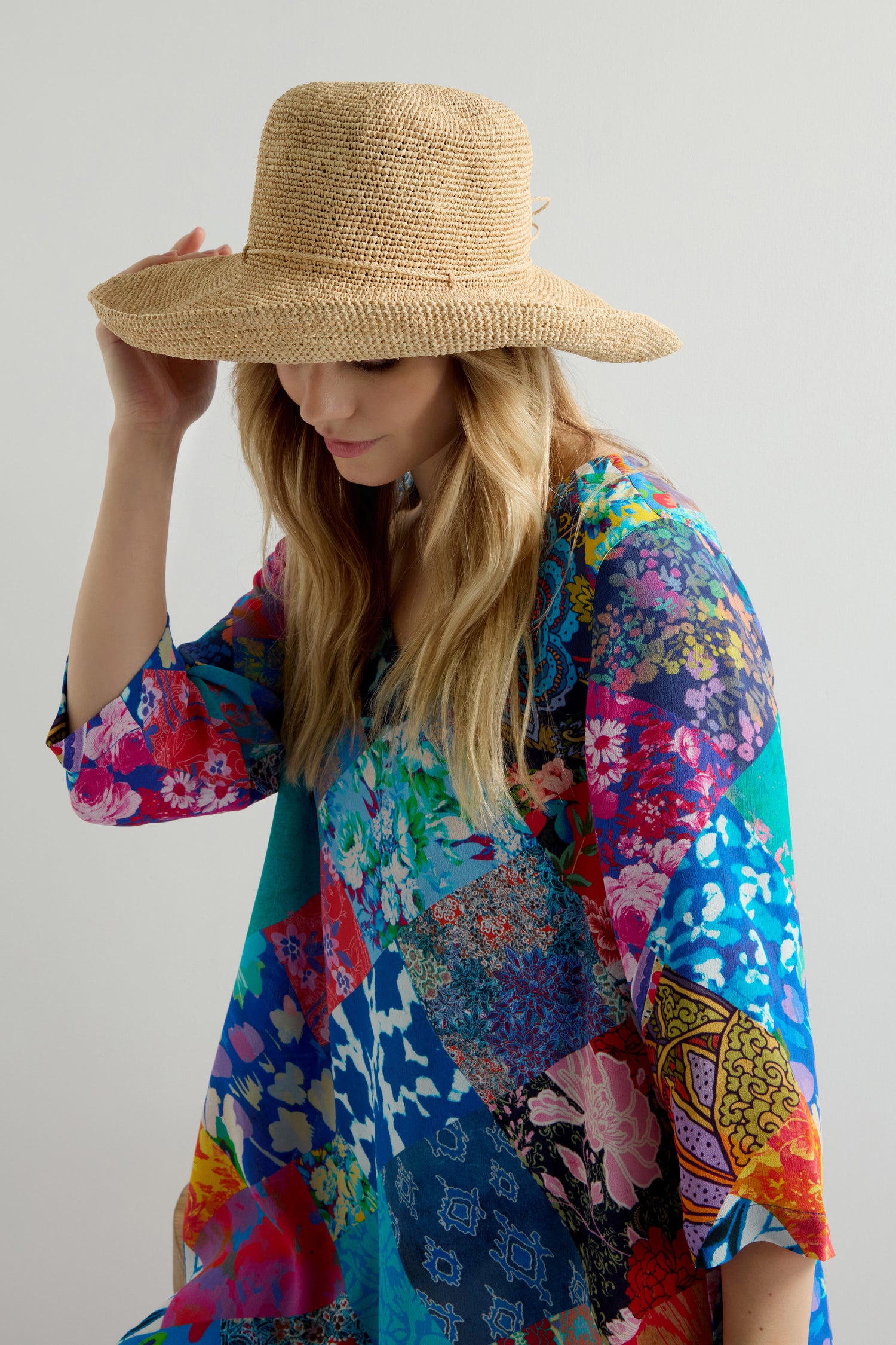 A person in a colorful floral dress tilts a Handwoven Wide Brim Raffia Hat with one hand, standing against a plain white background.