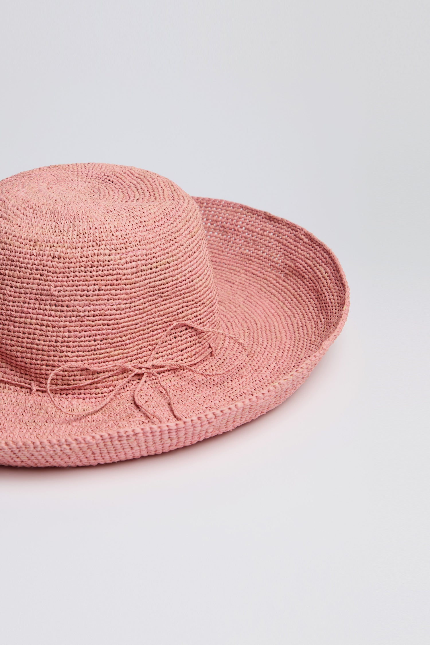The Handwoven Wide Brim Raffia Hat in pink, featuring a ribbon detail, rests stylishly on a white surface, providing sun protection.