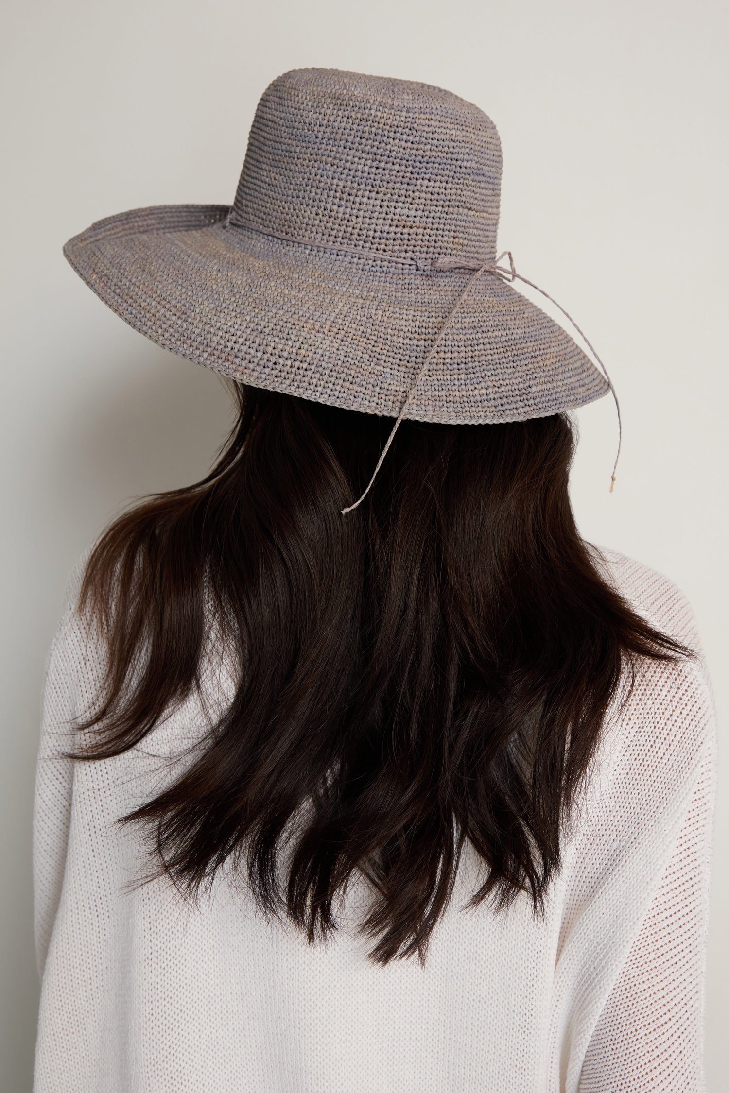 A person with long dark hair is shown from behind, wearing the Handwoven Wide Brim Raffia Hat and a light-colored sweater against a plain background.