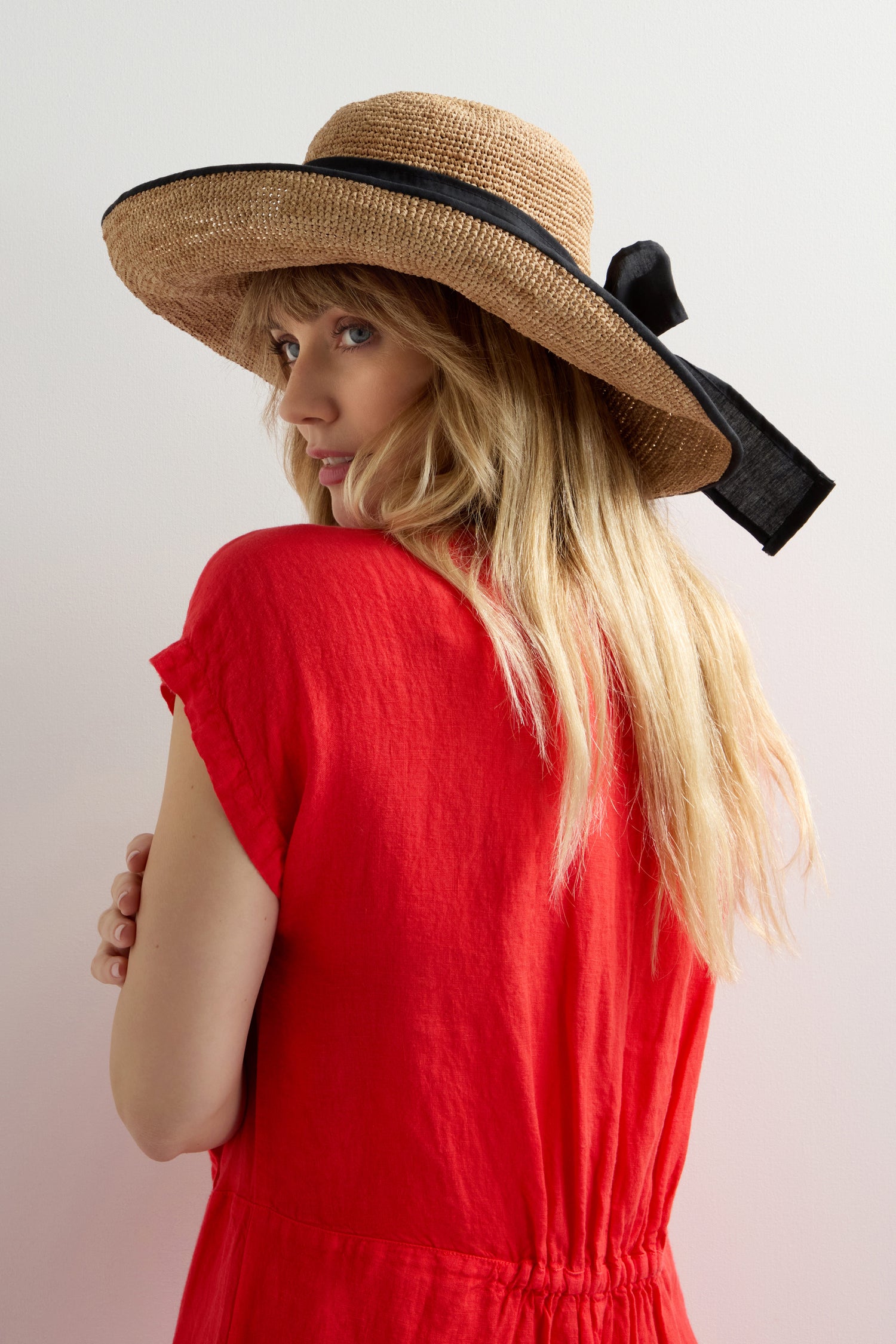 Wearing a red dress, a long-haired individual sports the Handwoven Ribbon Raffia Hat, glancing over their shoulder against a plain backdrop, effortlessly blending style and sun protection.