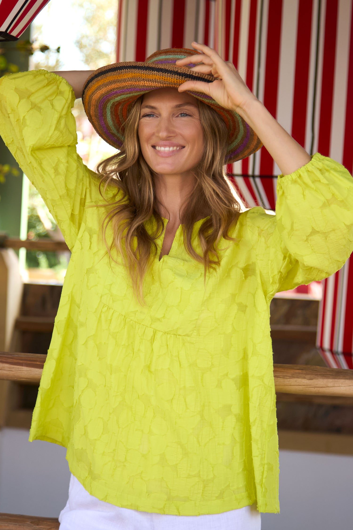 A woman in a yellow textured blouse and white pants smiles while holding the Handwoven Raffia Rainbow Stripe Hat, featuring a colorful wide brim, standing before red-and-white striped outdoor curtains.