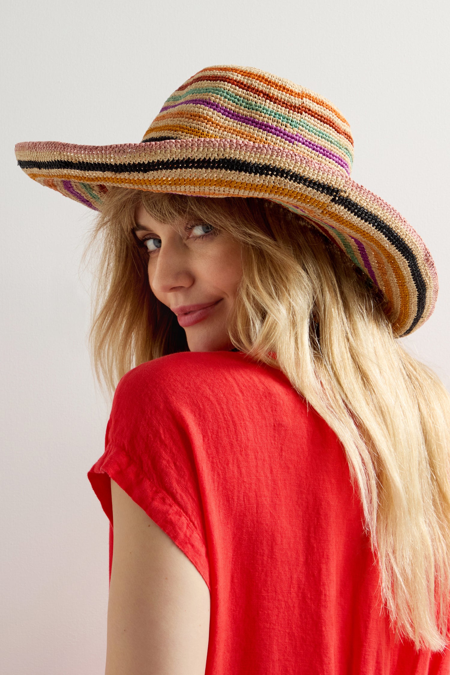 Wearing the Handwoven Rainbow Stripe Raffia Hat and a red shirt, a person exudes boho-chic style, glancing over their shoulder with a slight smile to showcase artisanal charm.