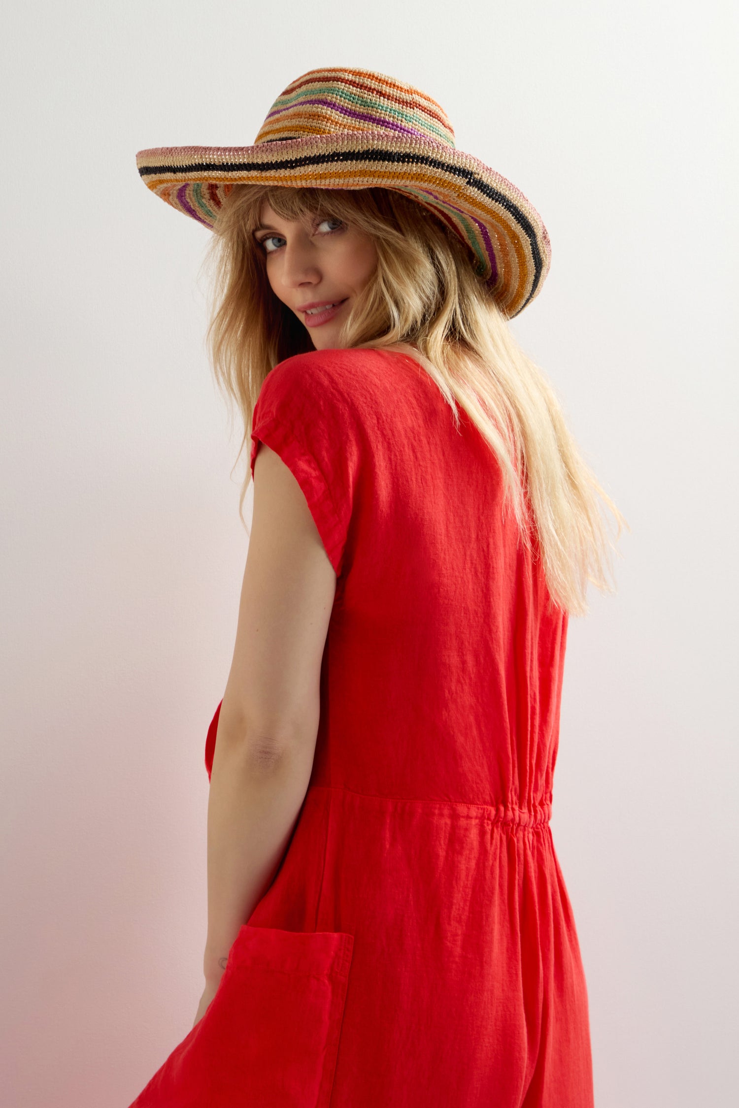 A person in a red dress, wearing a Handwoven Rainbow Stripe Raffia Hat, looks over their shoulder against a white backdrop, exuding artisanal charm.