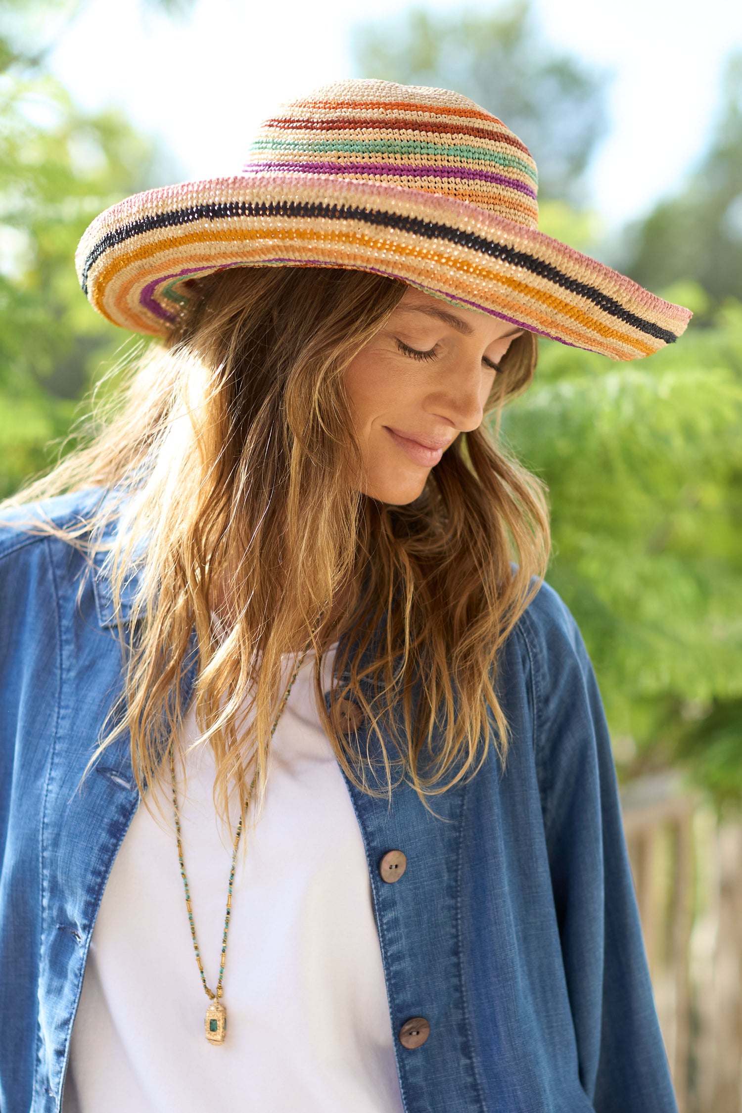 A woman outdoors, wearing a denim shirt and the Handwoven Rainbow Stripe Raffia Hat, gazes down with a slight smile.