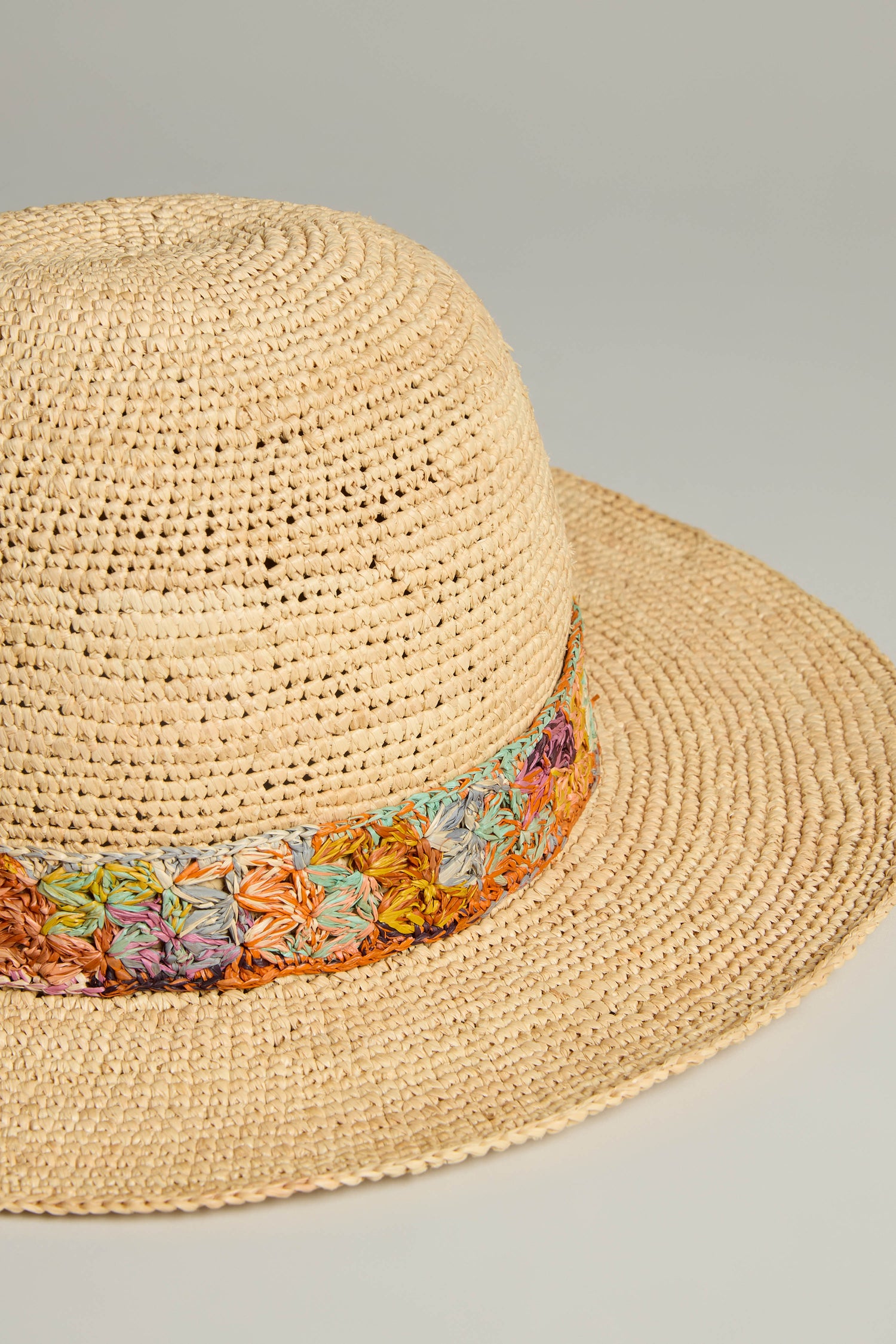 The Handwoven Flower Raffia Hat, with a vibrant multicolored band, offers sun protection and sustainable fashion against a light gray backdrop.