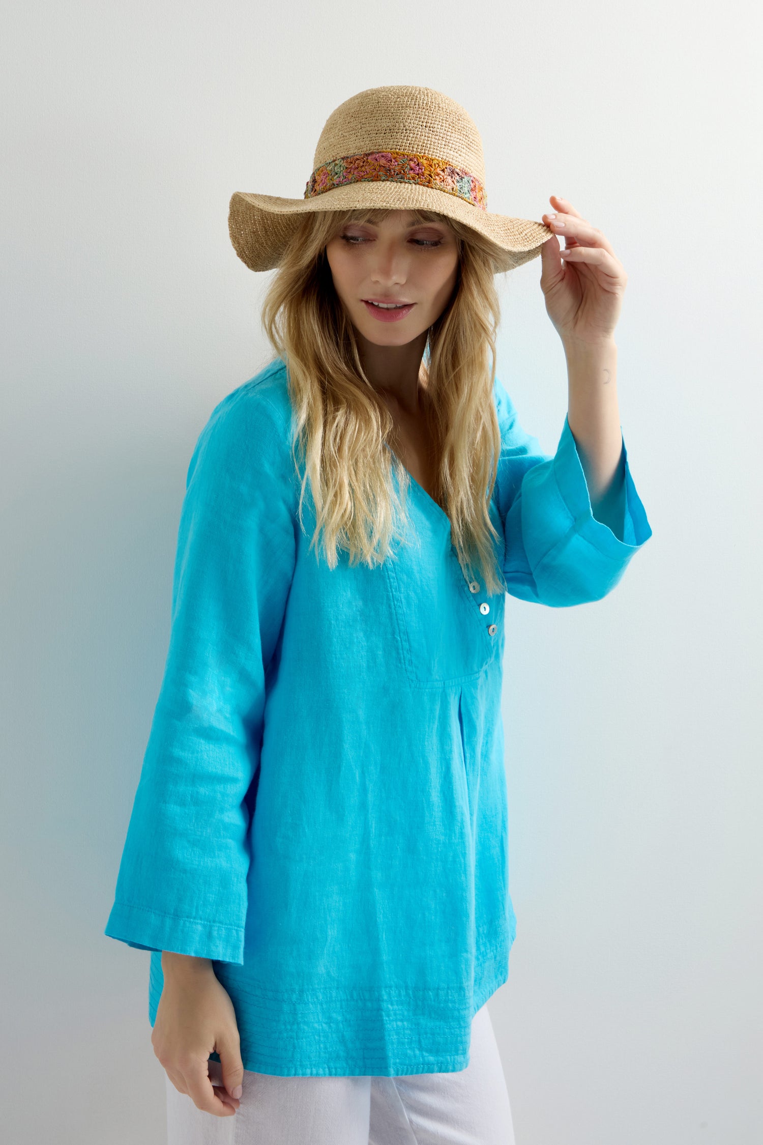 Wearing a blue tunic and the Handwoven Flower Raffia Hat with a vibrant band, someone stands against a light background, showcasing sustainable fashion and sun protection.