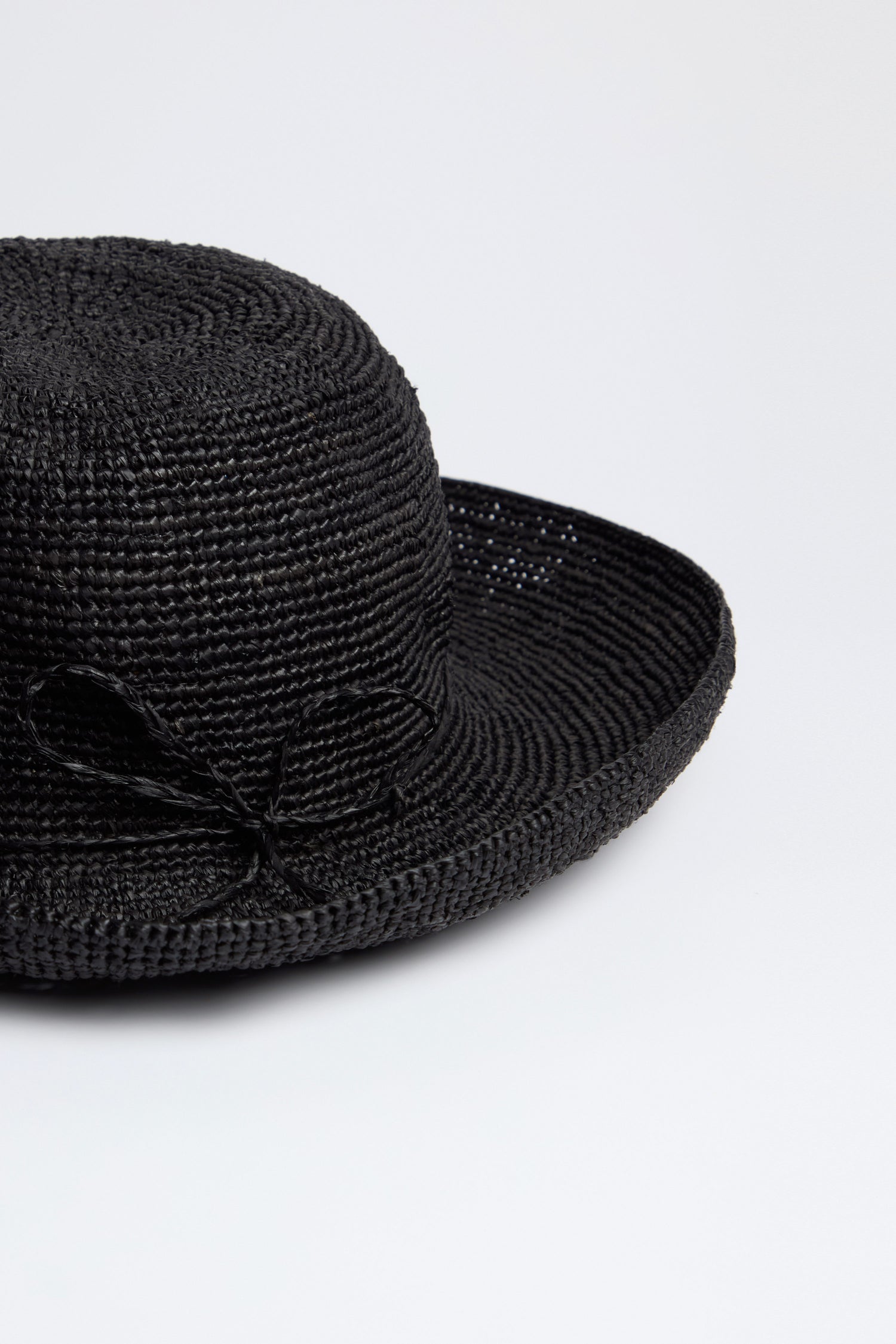 The Handwoven Raffia Hat, made with Madagascar techniques, has a black woven straw with a round crown and side bow. Set against a plain white background, this lightweight and breathable hat blends elegance and tradition.